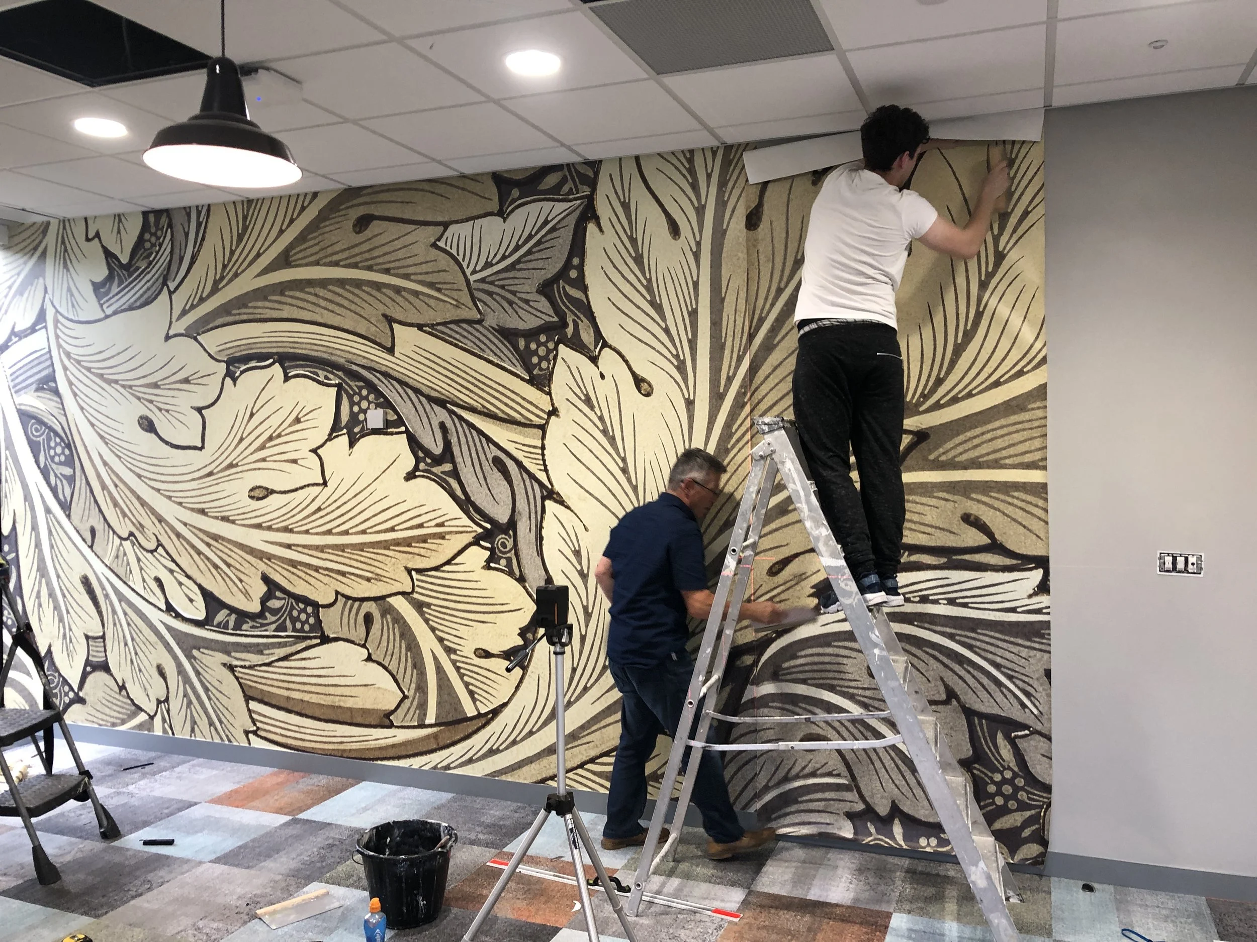 Two men installing a large decorative mural on an office wall, with one standing on a ladder and the other on the floor.