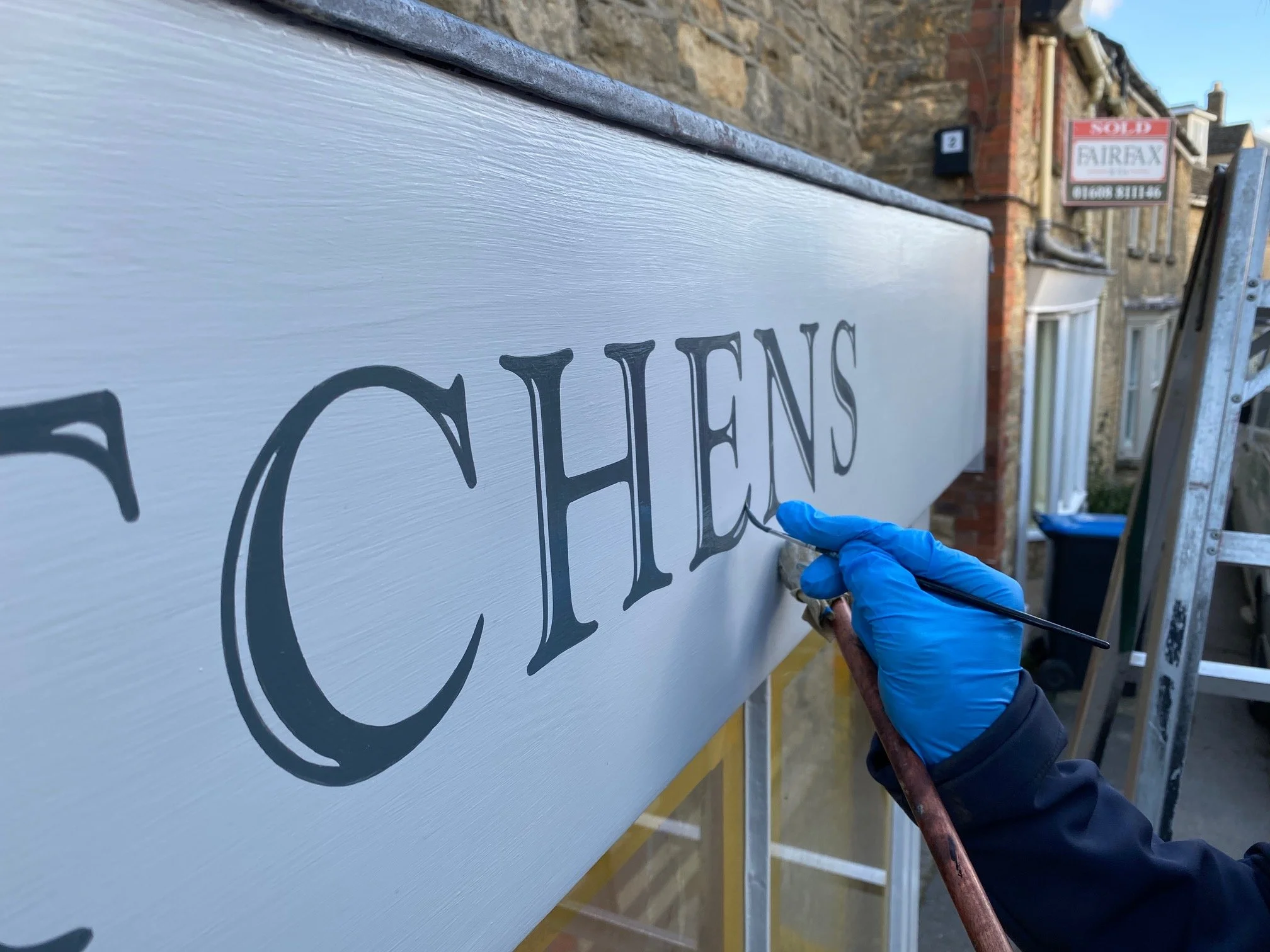 A person wearing a blue glove is painting the black lettering on a white sign with the word 'CHE'. A ladder and other buildings are visible in the background.
