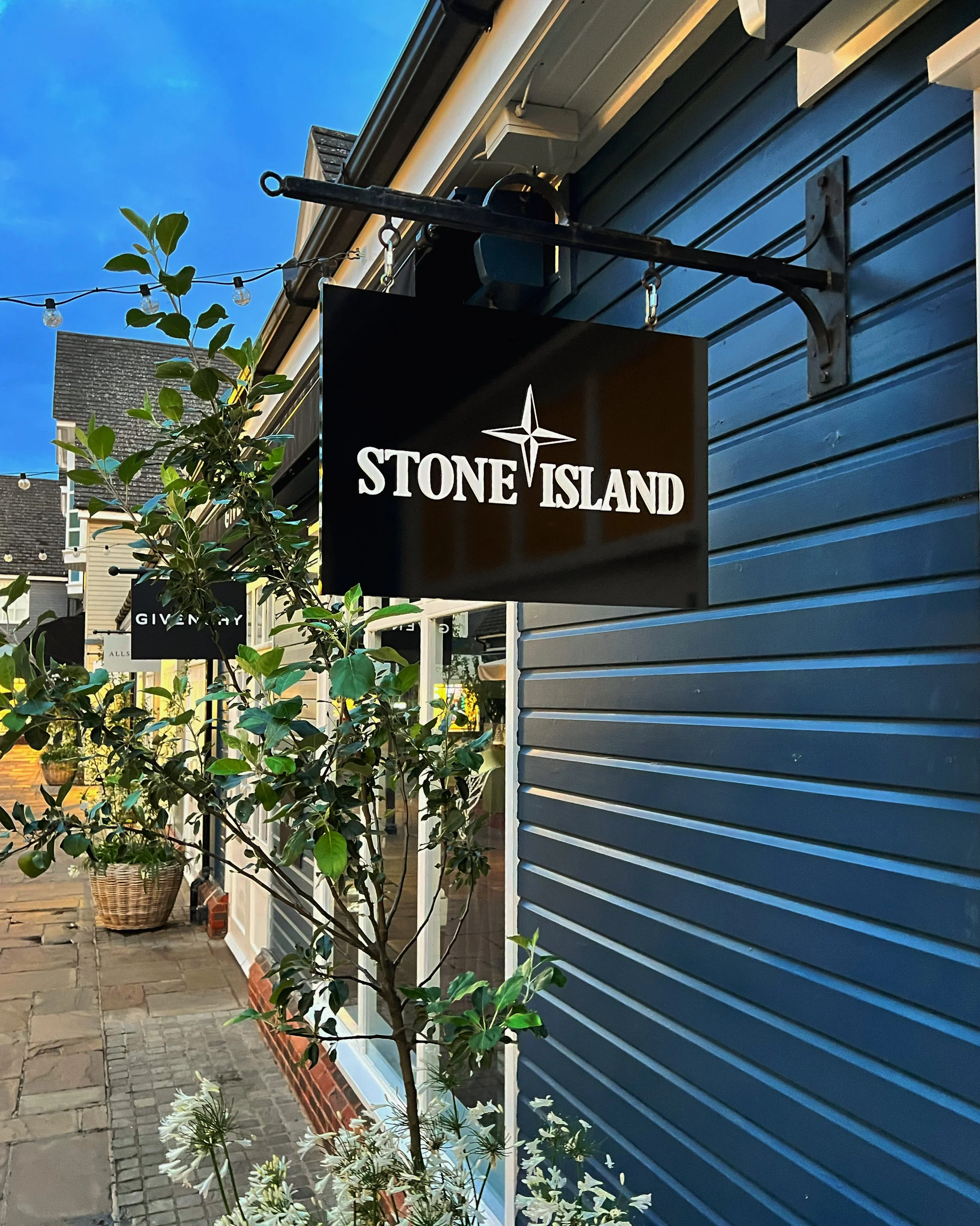 Stone Island store sign outside a blue building with outdoor plants and other store signs visible.