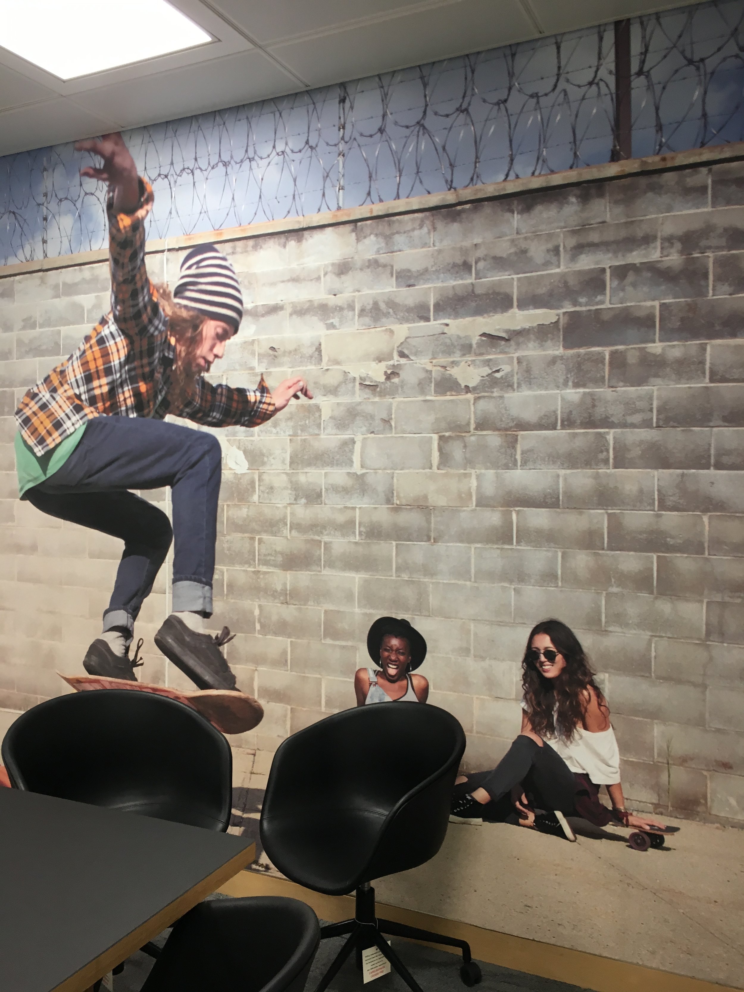 A large mural of three people, one skateboarder jumping in mid-air and two women on the ground laughing, on a brick wall in an office space.