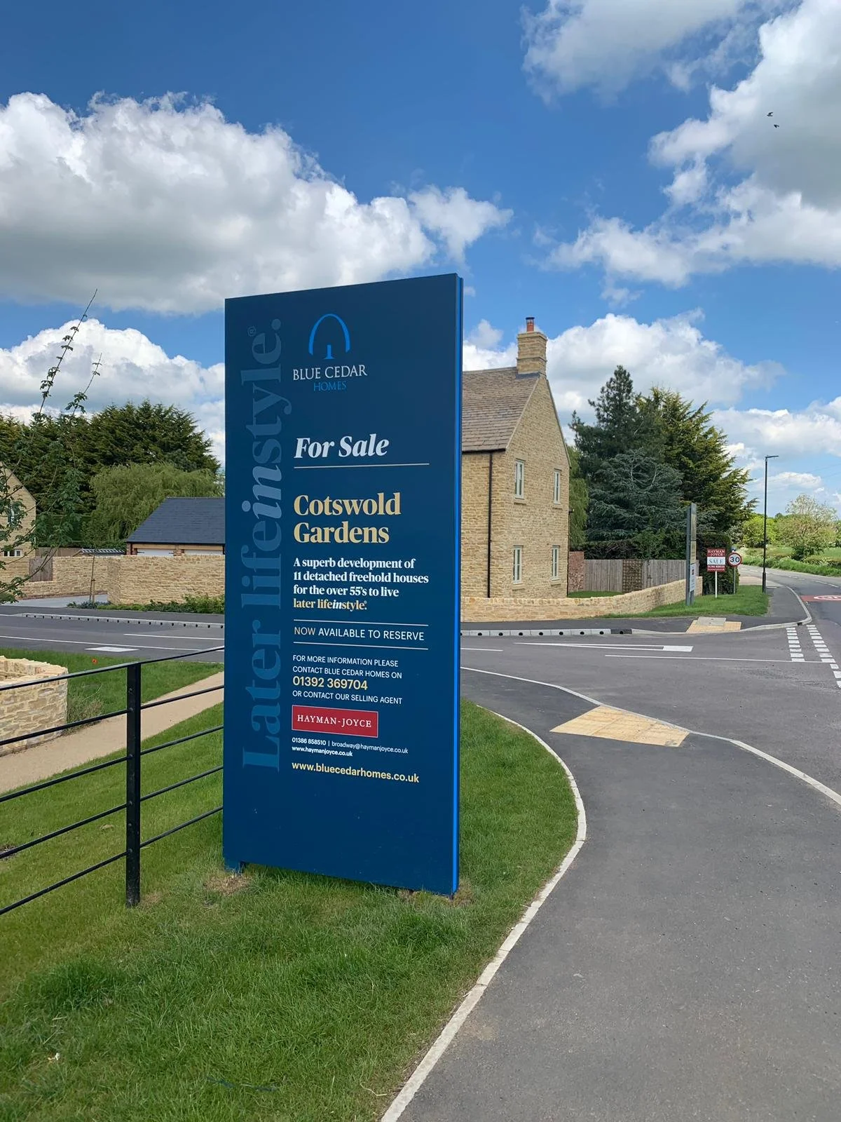 Blue sign advertising homes for sale at Cotswold Gardens, developed by Blue Cedar Homes, located near a sidewalk with a brick house, green trees, and a partly cloudy sky in the background.