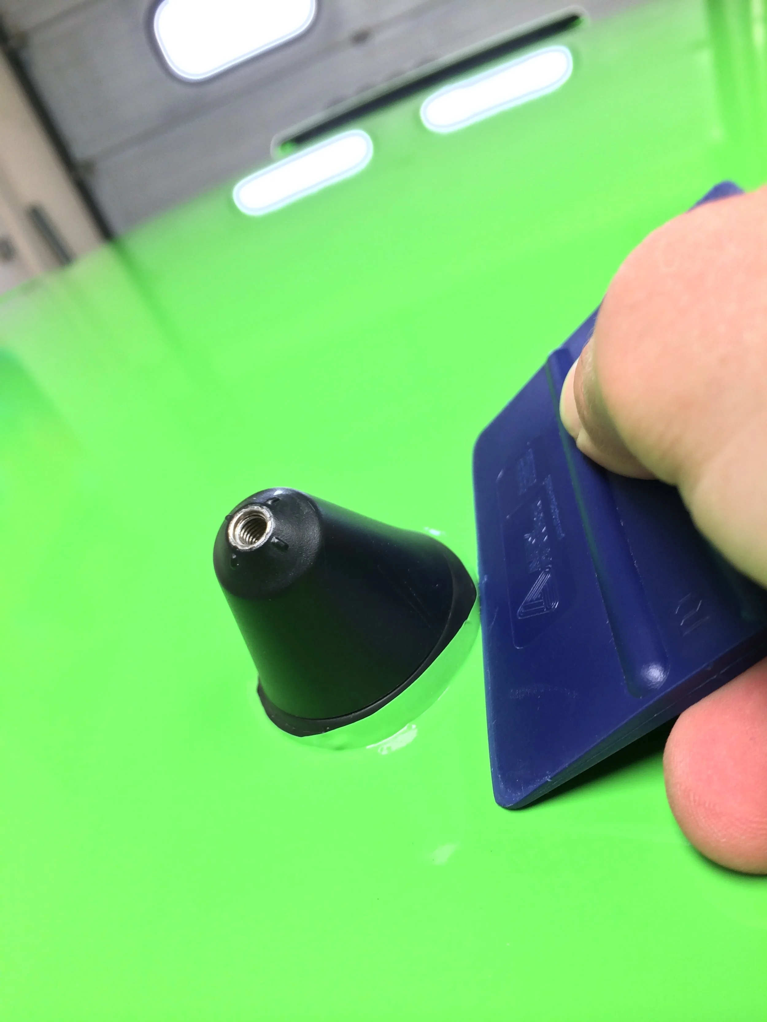 A person's hand holding a blue device near a black cone-shaped object on a bright green surface.