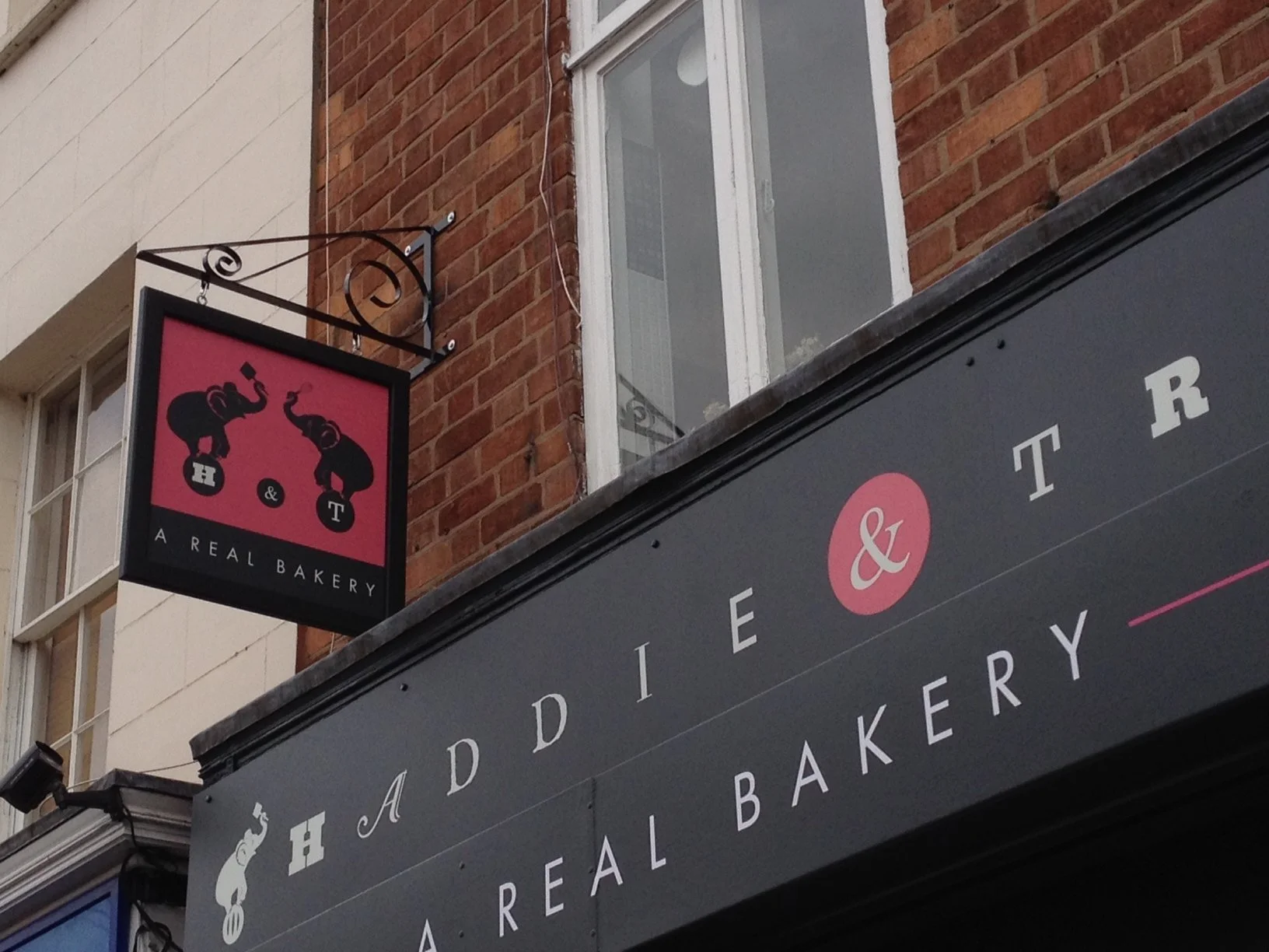 Sign of a bakery with two elephants, one holding a bread loaf and the other a whisk, on a red background. The sign also says 'A Real Bakery'.