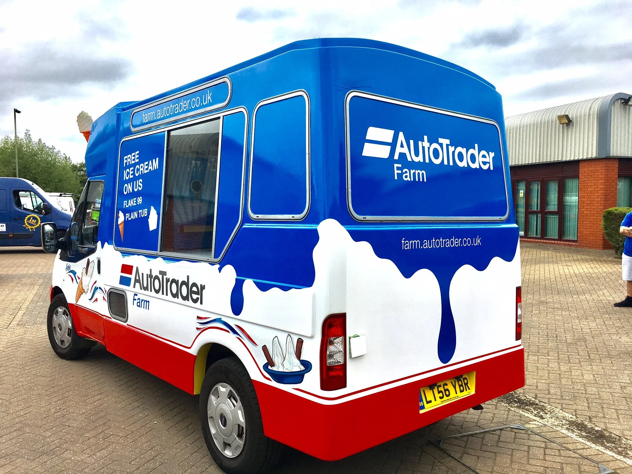 A colorful ice cream truck with branding for AutoTrader Farm, featuring blue, red, and white graphics including ice cream cones and a bowl of ice cream, parked on a paved lot.