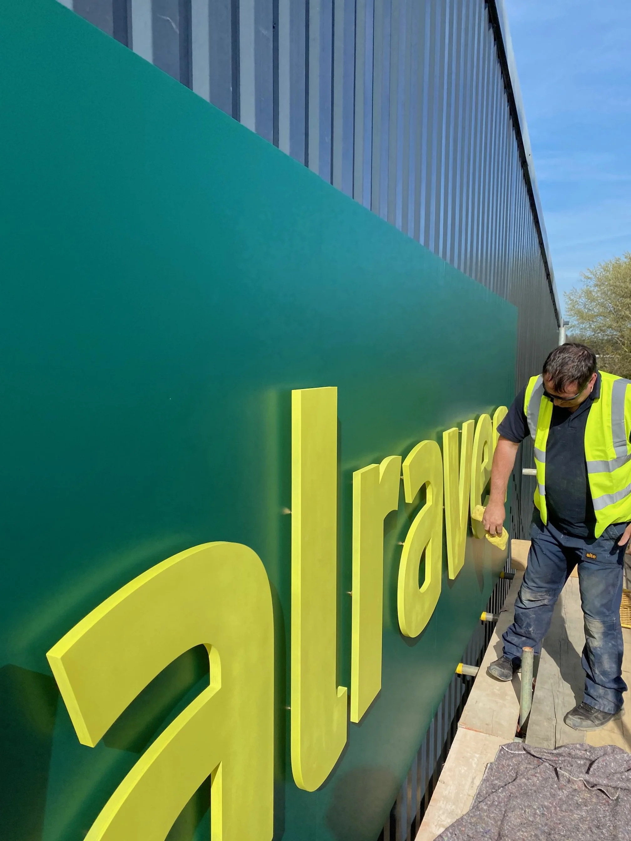 A worker cleaning a large green sign with yellow letters outside a building on a sunny day.