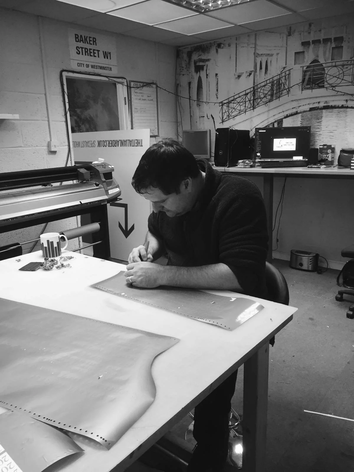 A man sitting at a work table, measuring fabric, surrounded by tools and equipment in a workshop with posters and monitors in the background.