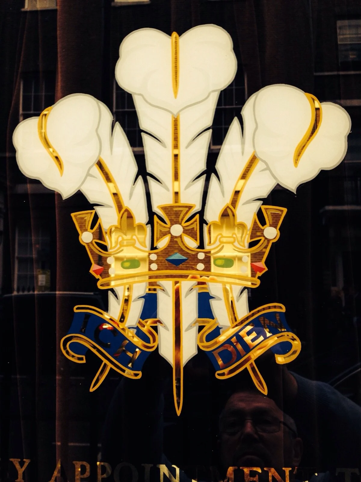 A stained glass window featuring a shield with a crown, three white feathers, and a blue ribbon with yellow text that reads 'CHILDREN DIED'.