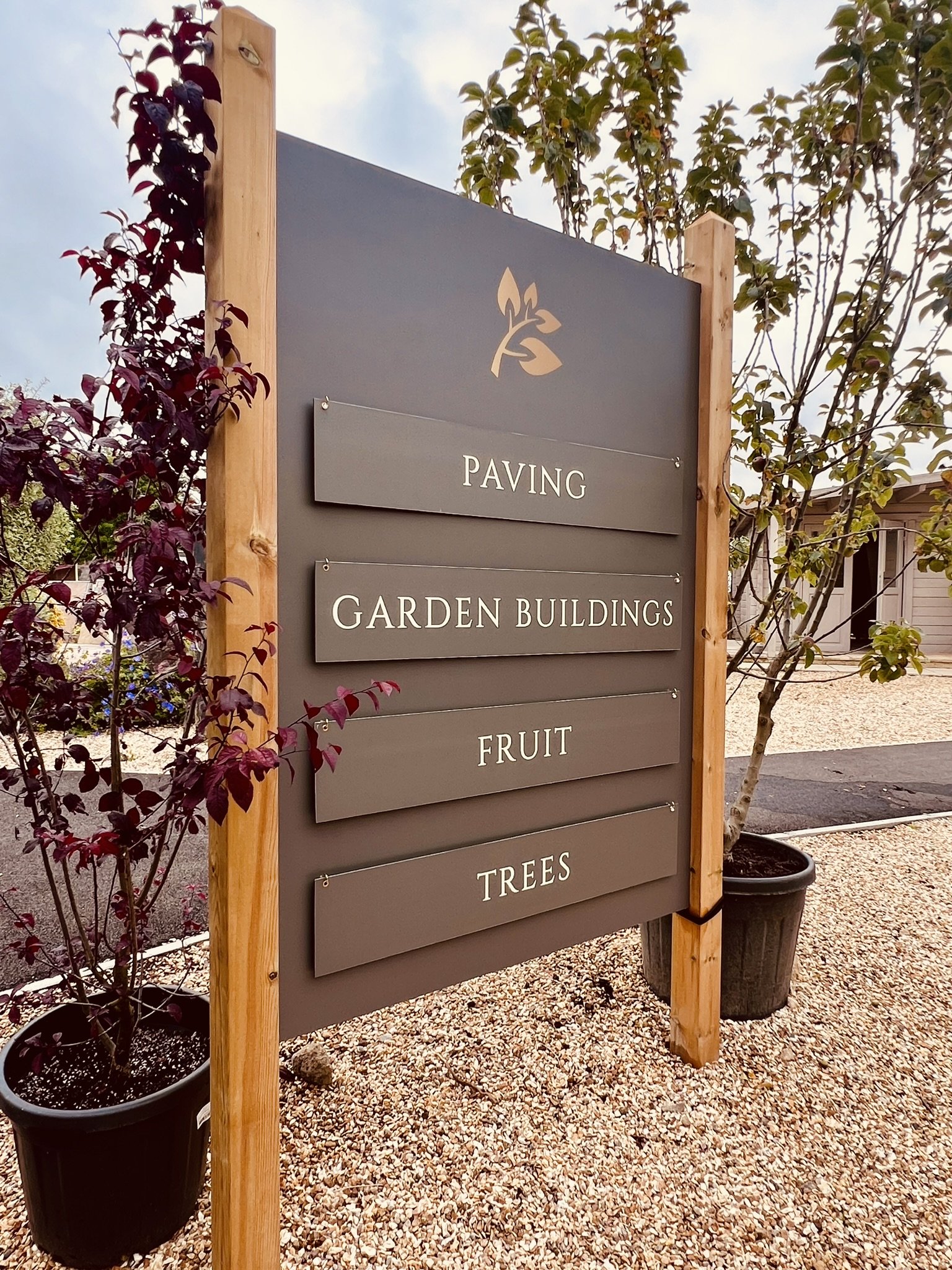 Signboard at a garden center listing directions: Paving, Garden Buildings, Fruit, Trees, with potted plants nearby and gravel ground.