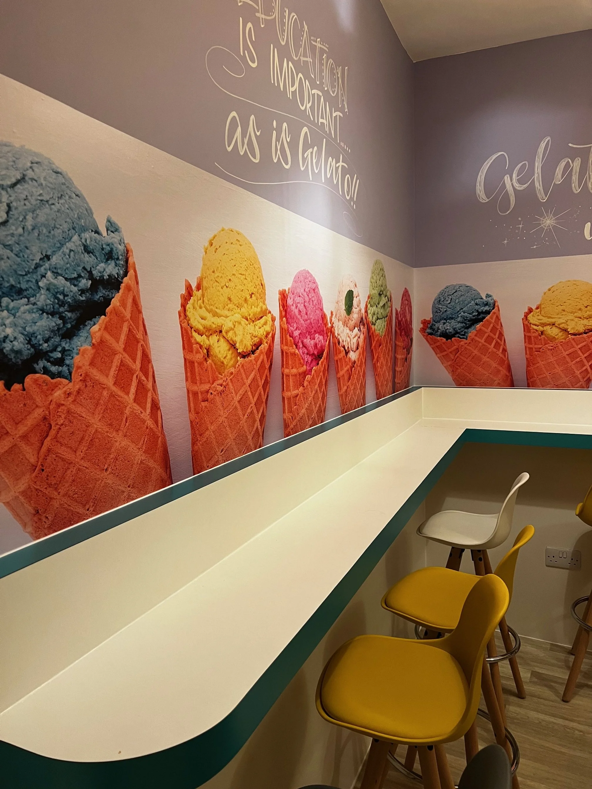 Decorative wall in an ice cream shop with colorful ice cream cones and a quote about ice cream. The wall features images of different flavored ice creams in waffle cones, and there are yellow and white chairs at a white counter.