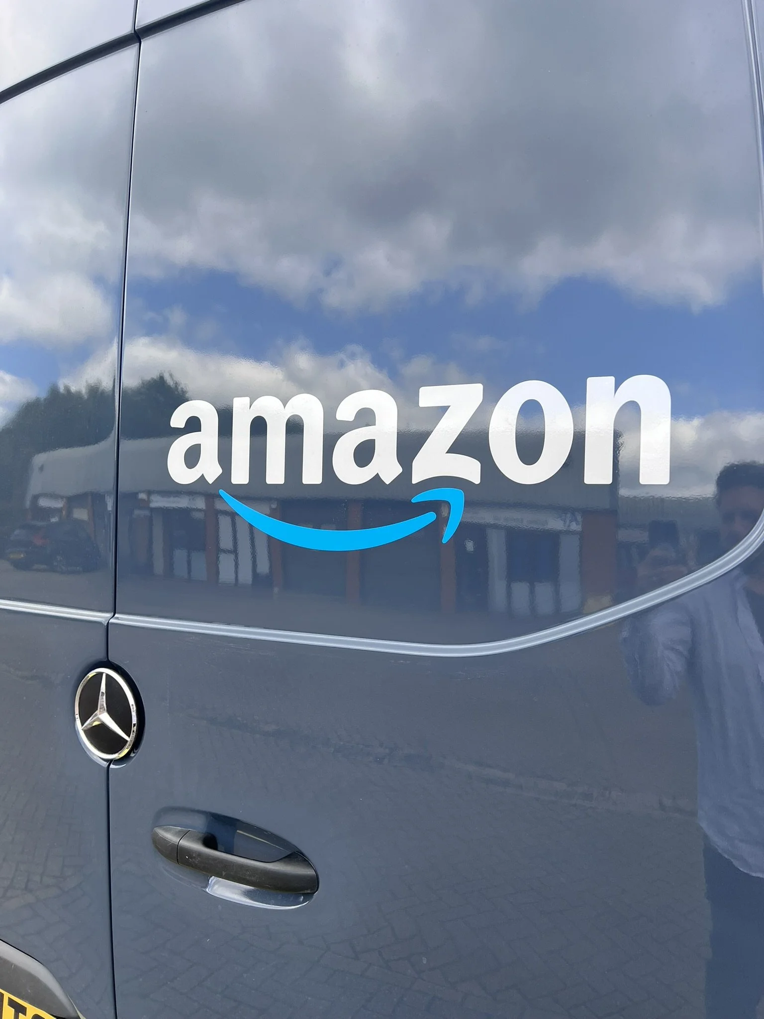 A Mercedes-Benz van with an Amazon logo sticker on the side reflecting the sky and clouds.