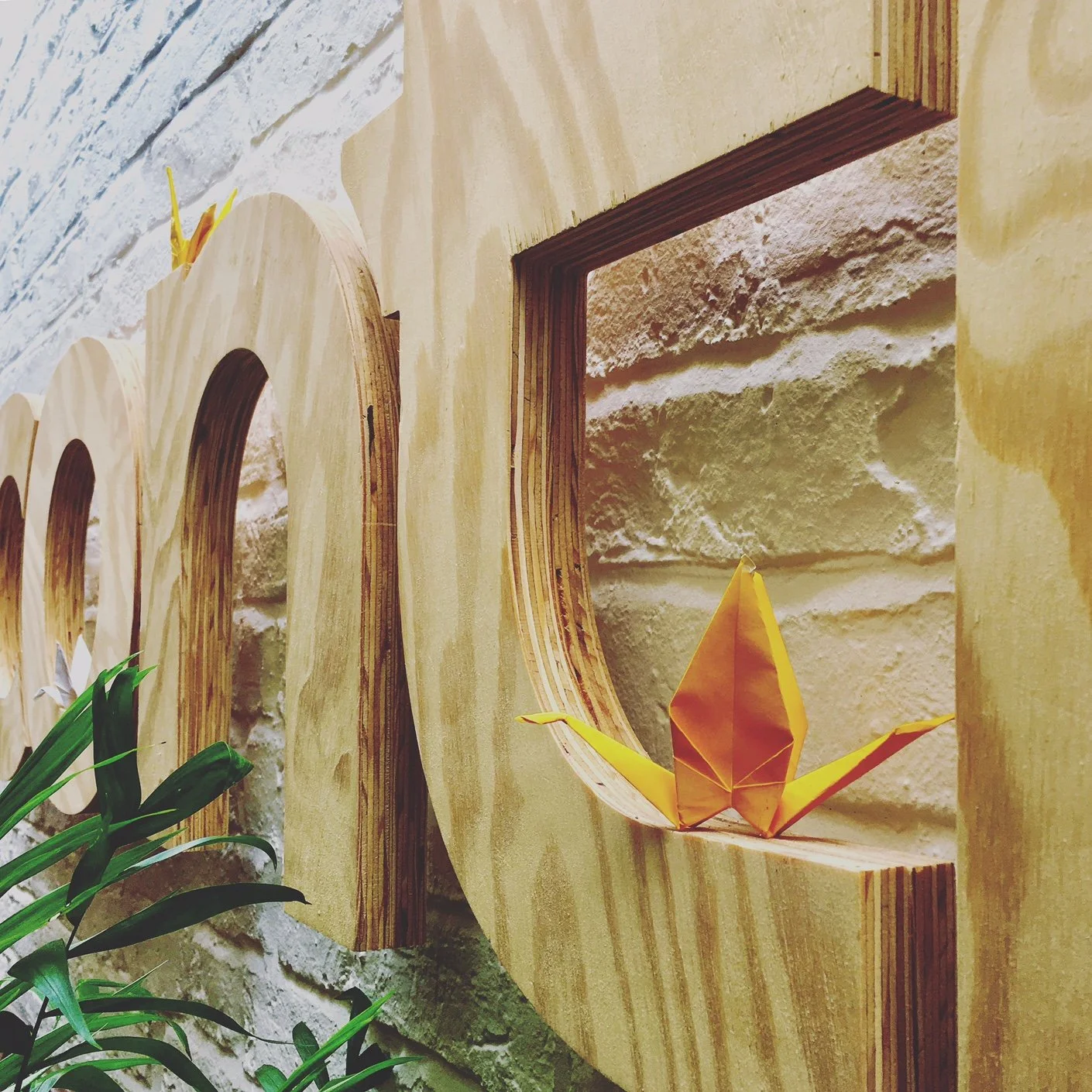 A close-up of a wooden wall decor with a cutout window, featuring an orange origami paper crane resting on the wooden frame, with some green plants in the foreground.