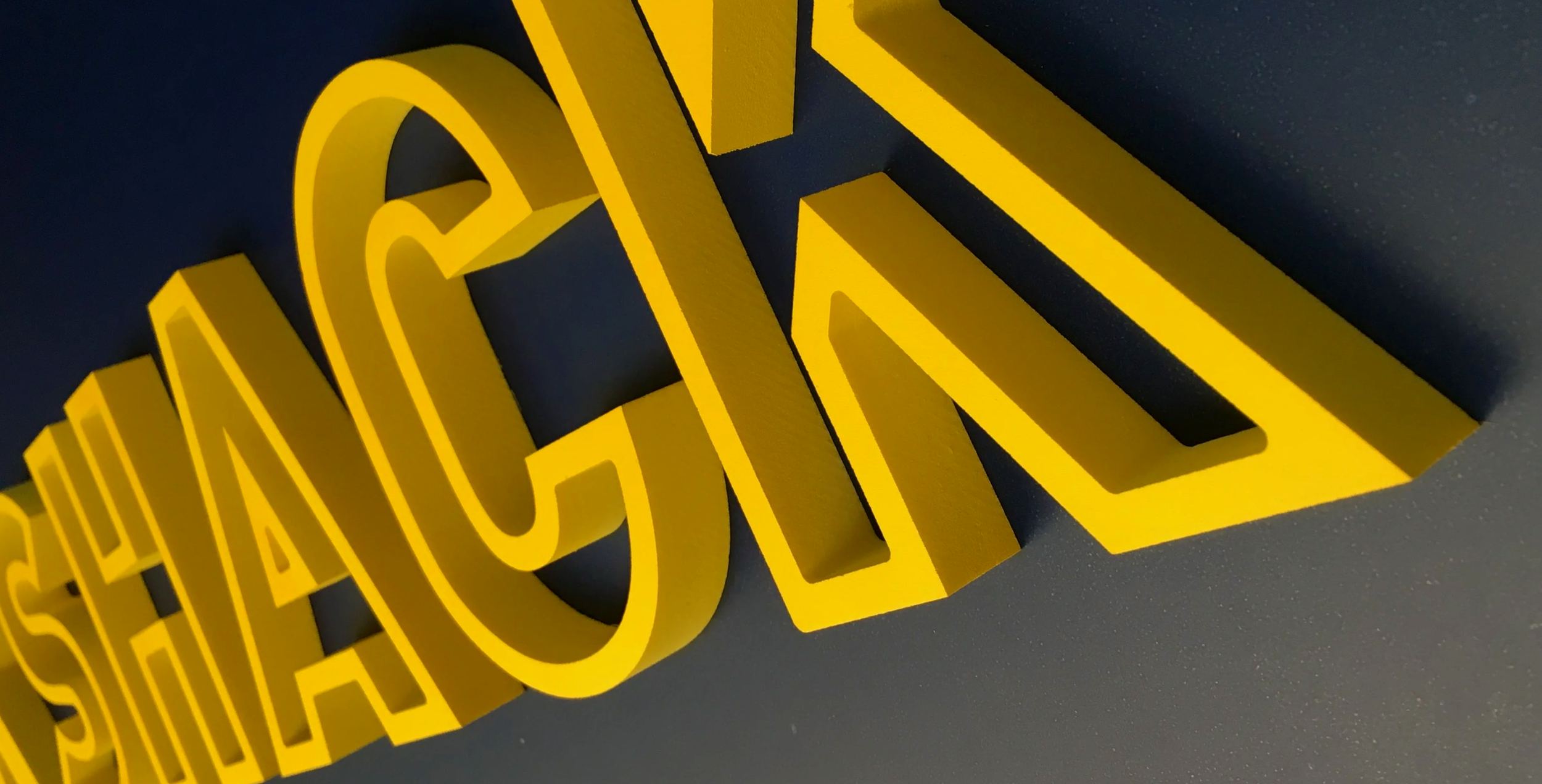 Close-up of yellow three-dimensional logo letters on a dark grey background.