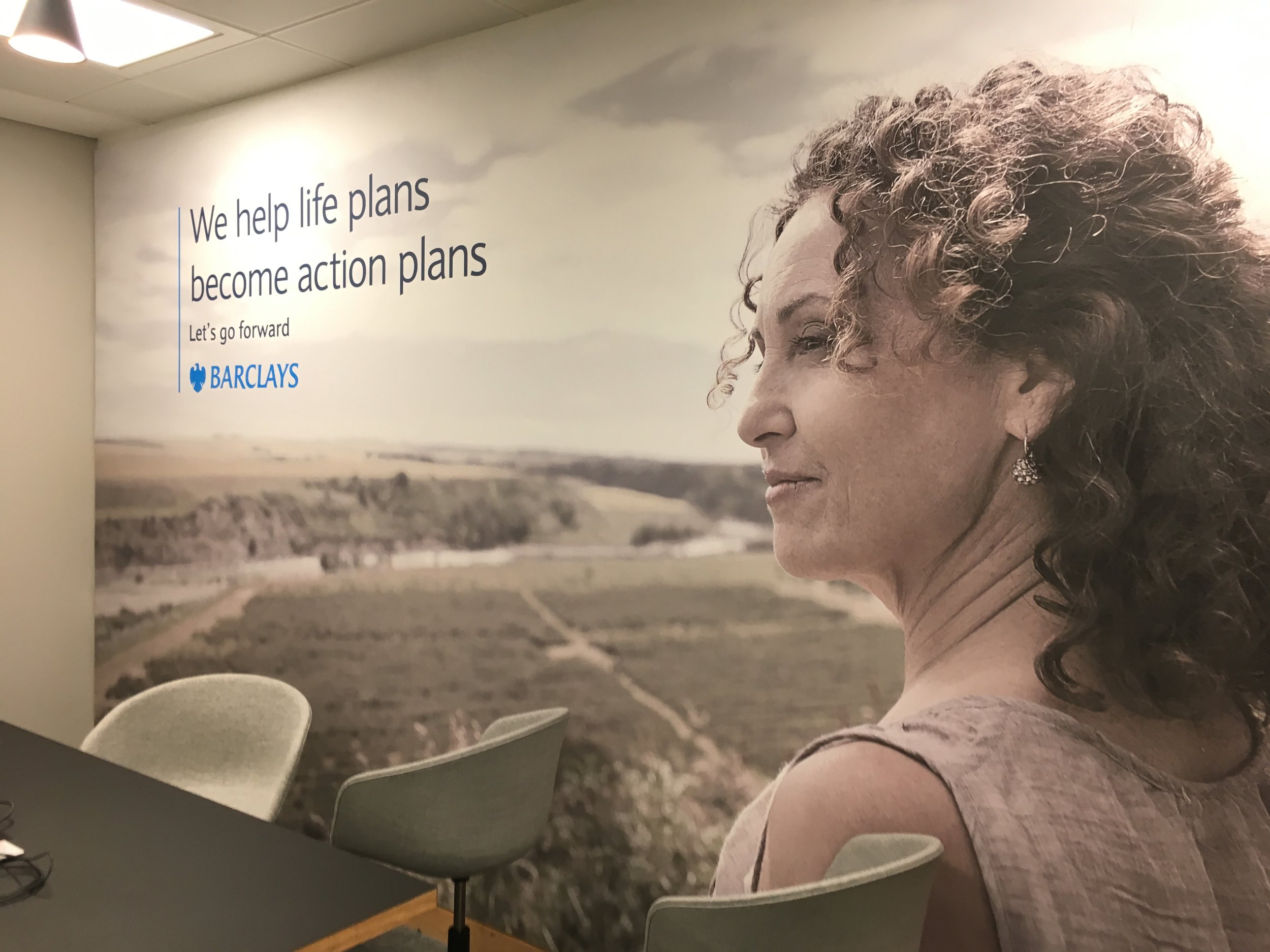 Office wall with large black and white mural of a woman with curly hair, sitting at a conference table, with a quote about helping life plans become action plans and the Barclays logo.