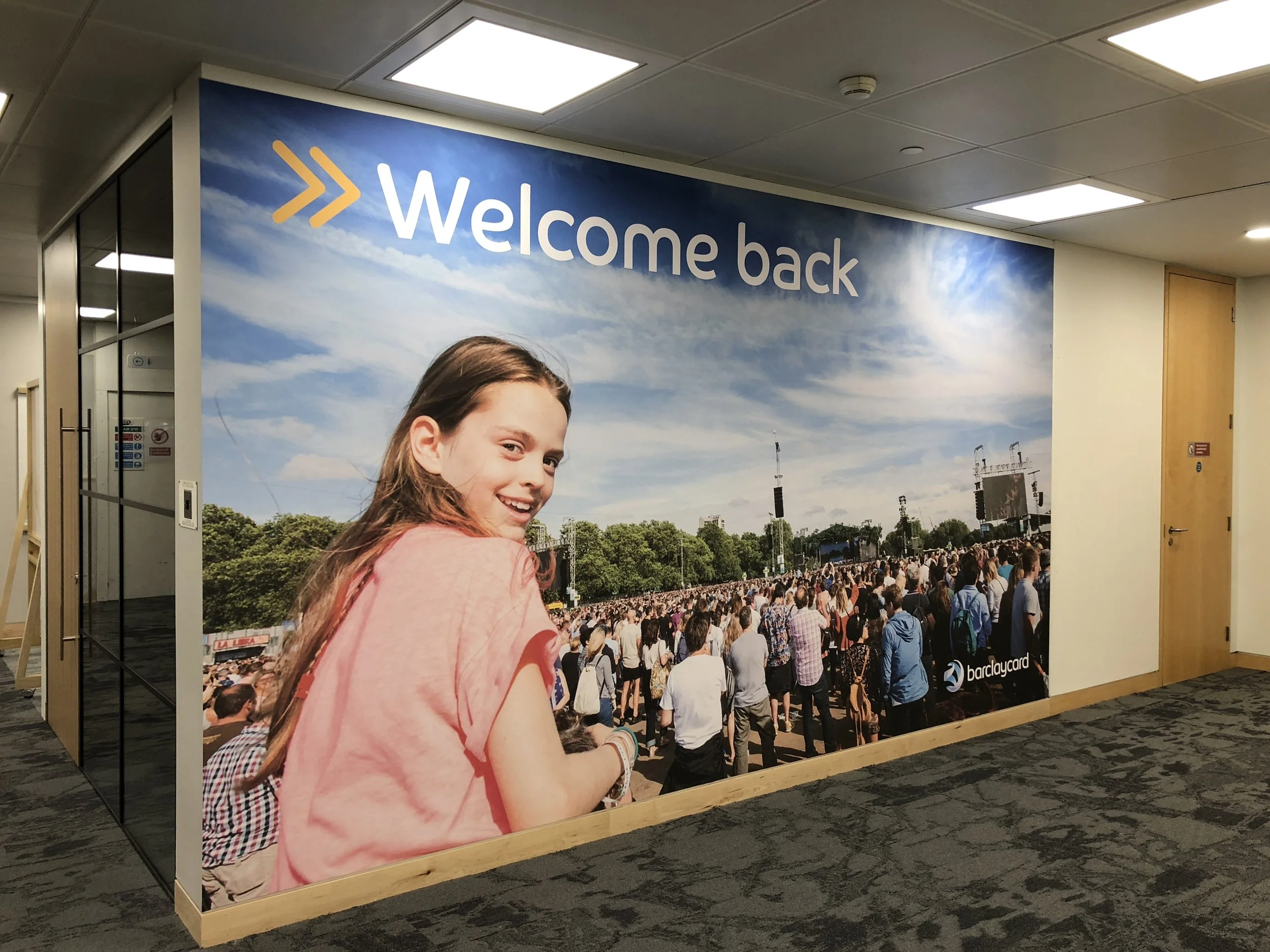 Large indoor wall mural with a young girl smiling and looking over her shoulder, crowds of people at an outdoor event, and the text 'Welcome back' at the top. The mural includes the Barclaycard logo.