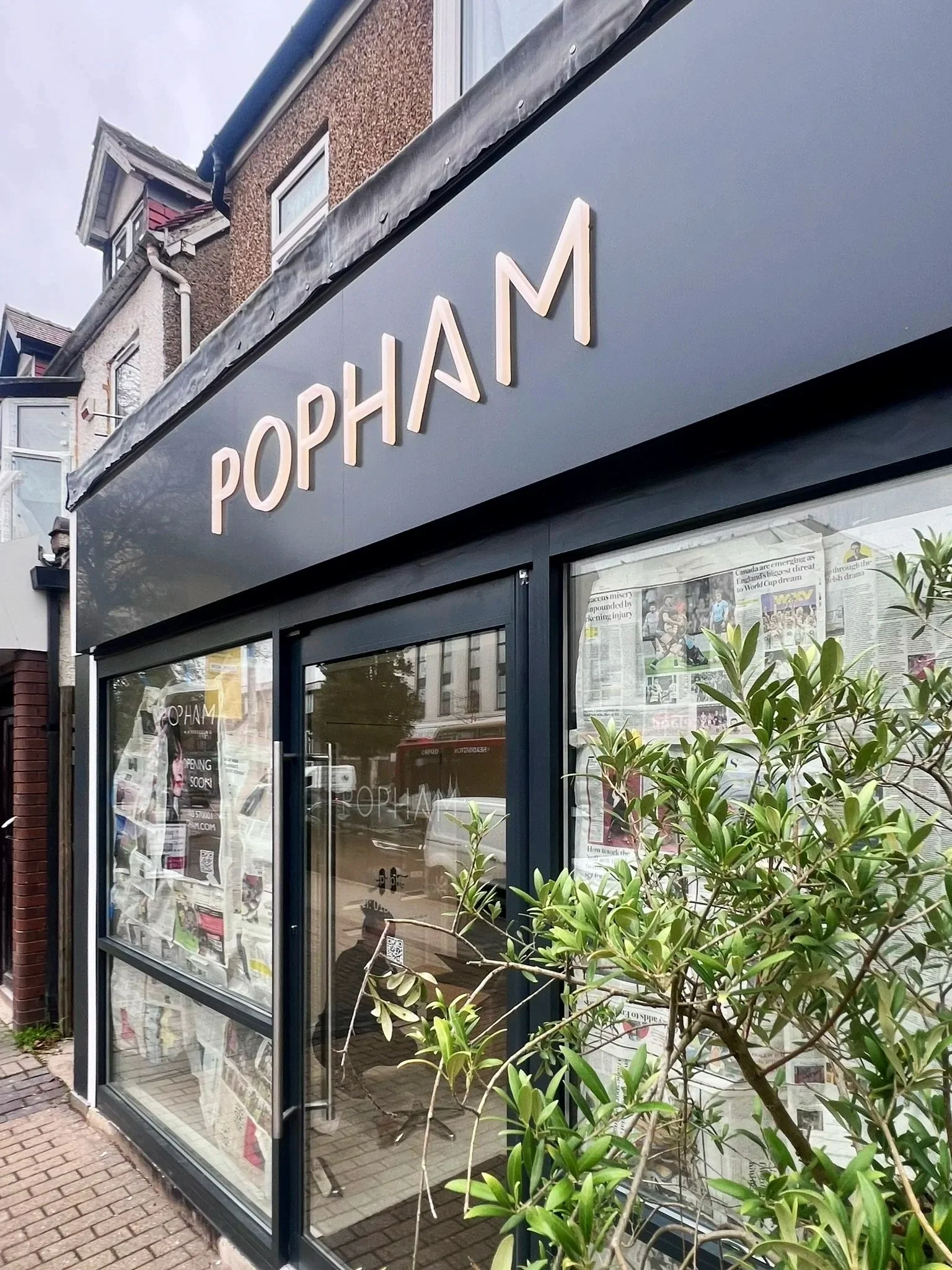 Exterior view of a storefront with the sign 'POP HAM' above the entrance, with newspaper-covered windows and some greenery in front.
