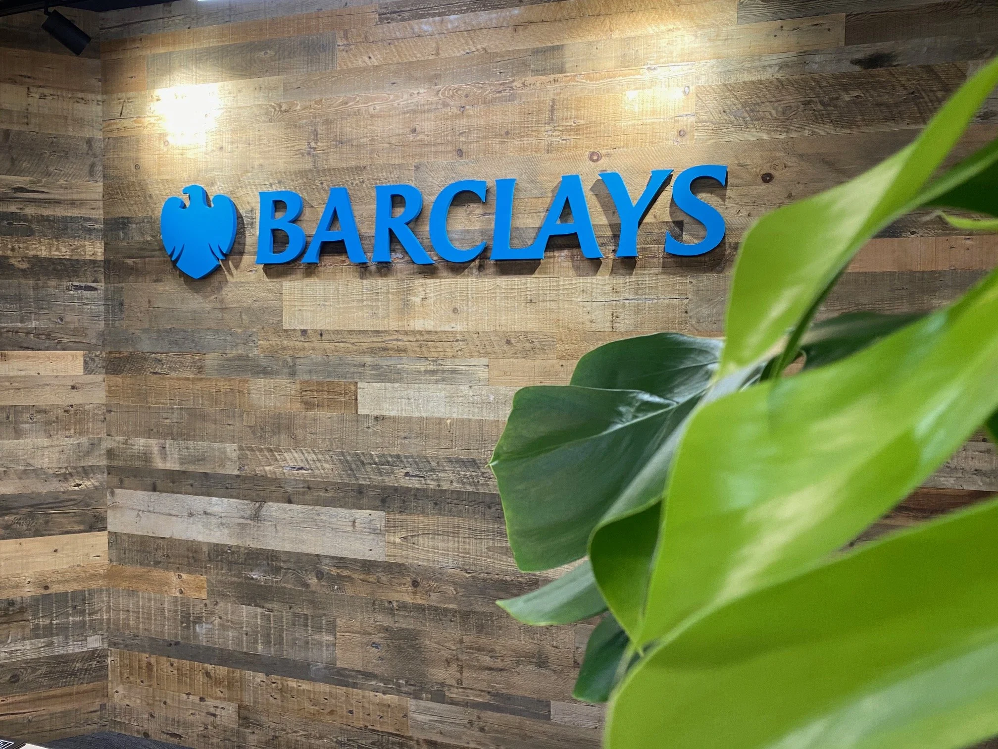 Barclays logo on a wood-paneled wall with green plant leaves in the foreground.