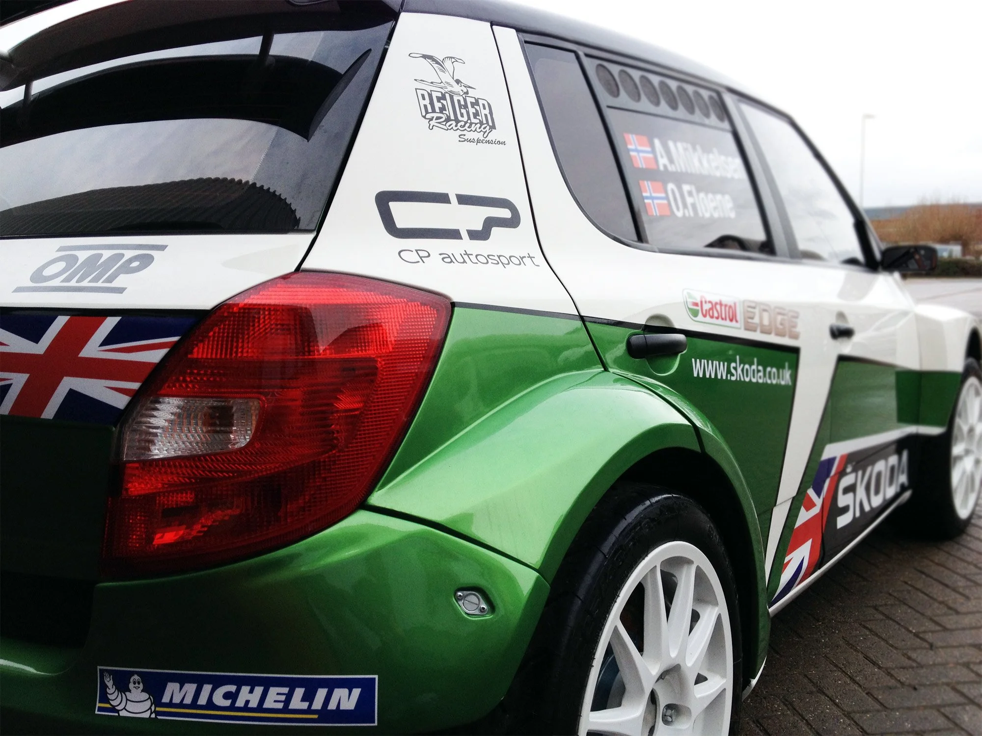 Close-up of a rally car with green, white, and black decals, displaying various sponsor logos and the names of drivers on the side window.