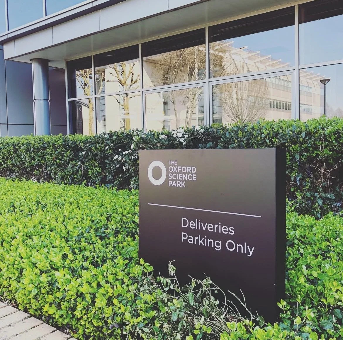 Sign indicating deliveries parking only outside a modern building with glass windows surrounded by green bushes.