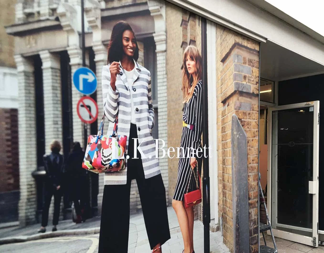 Large advertisement featuring two women in stylish black and white striped outfits on a city street; one woman has a colorful patterned handbag, the other holds a small red purse; behind them is a brick building and a street sign.