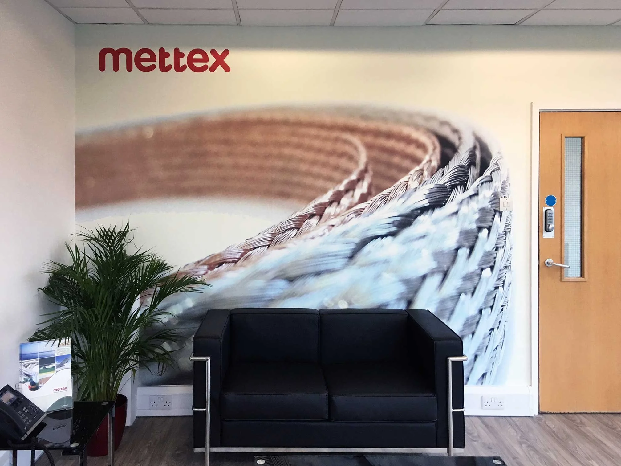 A black sofa, a green potted plant, and a small table with brochures and a telephone in a lobby with a large wall mural of woven baskets and the word 'mettex' in red letters.