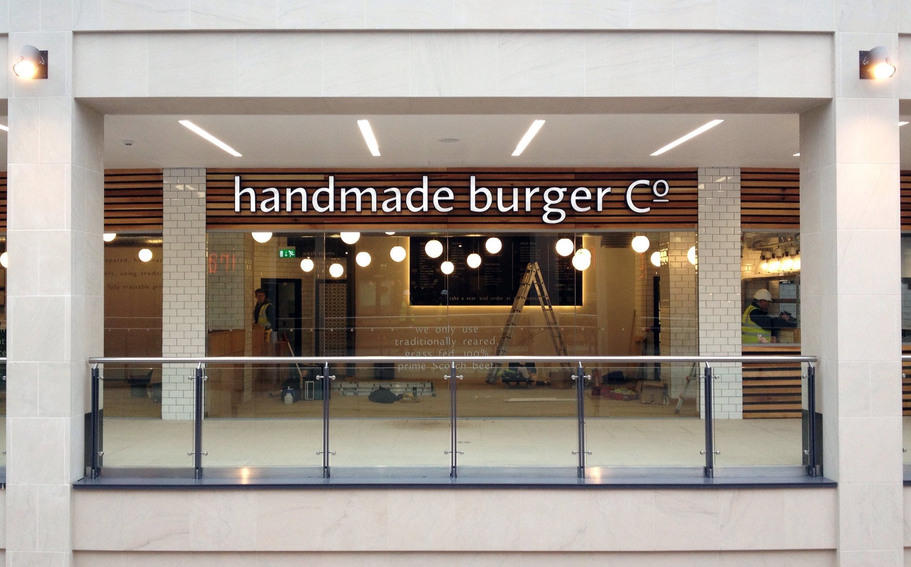 Front of a restaurant named handmade burger Co. with a glass facade, white brick columns, and wooden slat decorative panels. Inside, workers are seen with ladders, and hanging round lights are visible.