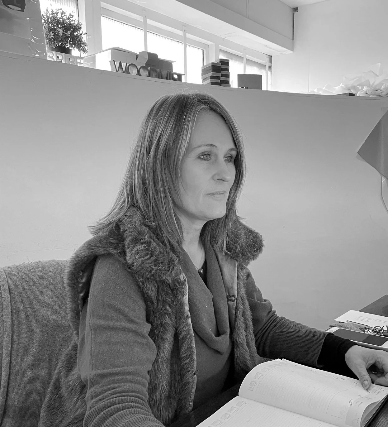 A woman with shoulder-length hair, sitting at a desk, looking to the side, with an open notebook and pen in front of her. The background shows modern office decor with windows and shelves.