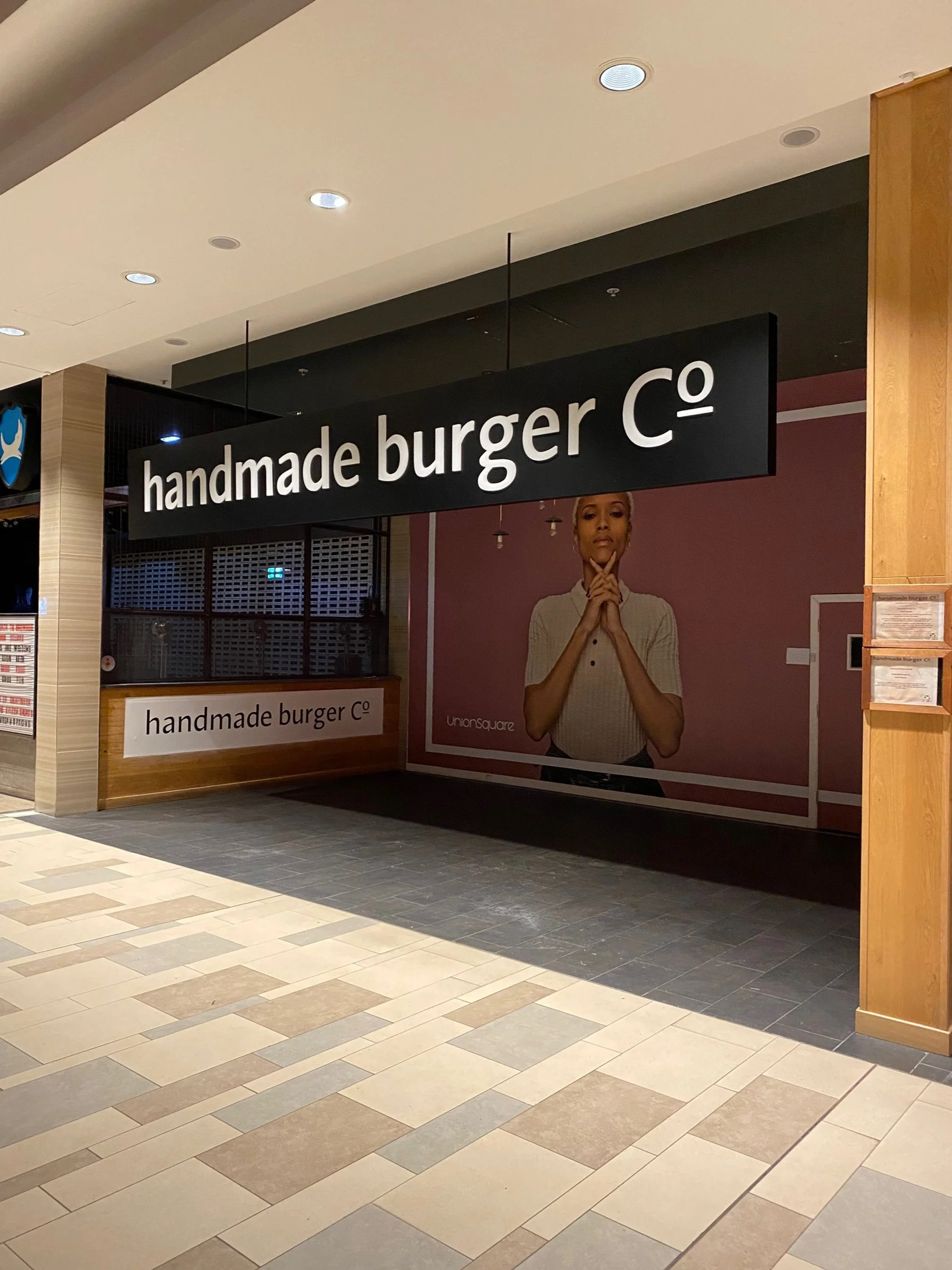 Storefront for 'handmade burger co' with a large sign hanging from the ceiling and a smaller sign on the counter. There is a large image of a woman with her hands under her chin in front of a pink background.