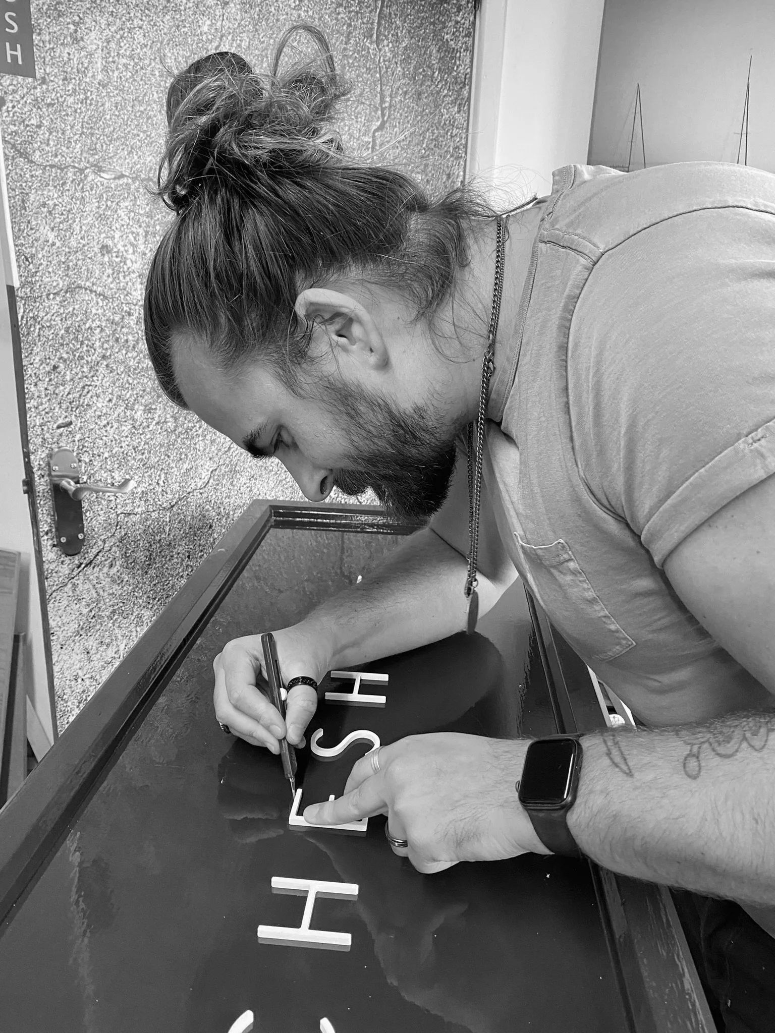 A man with a beard and long hair tied in a bun signs a sign with the text 'FISH' on a glossy surface. He is wearing a light-colored t-shirt, a watch, and a necklace, and appears focused on signing.