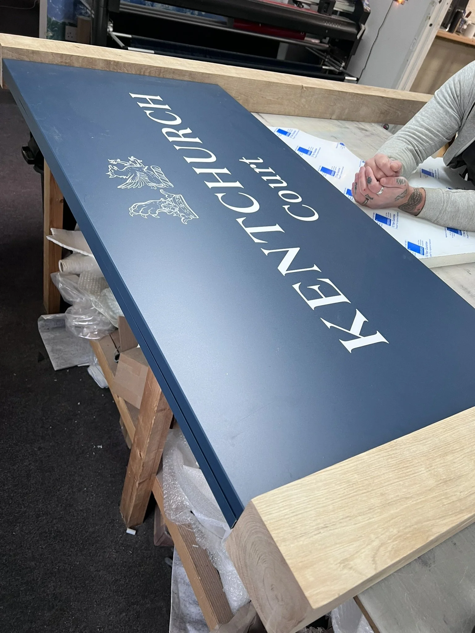A blue sign with white text reads "KENNETT CHURCH CO., LTD" with a decorative emblem. The sign is on a worktable in a workshop, with a person sitting nearby.