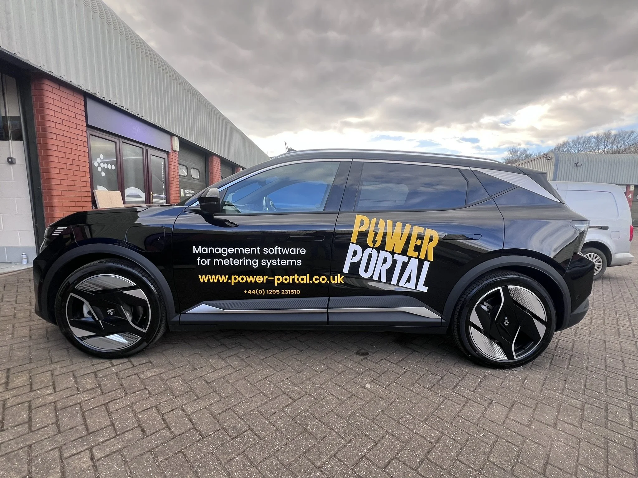 Black electric car parked outside a building with advertisement for Power Portal, a company offering management software for metering systems, with website and phone number displayed.