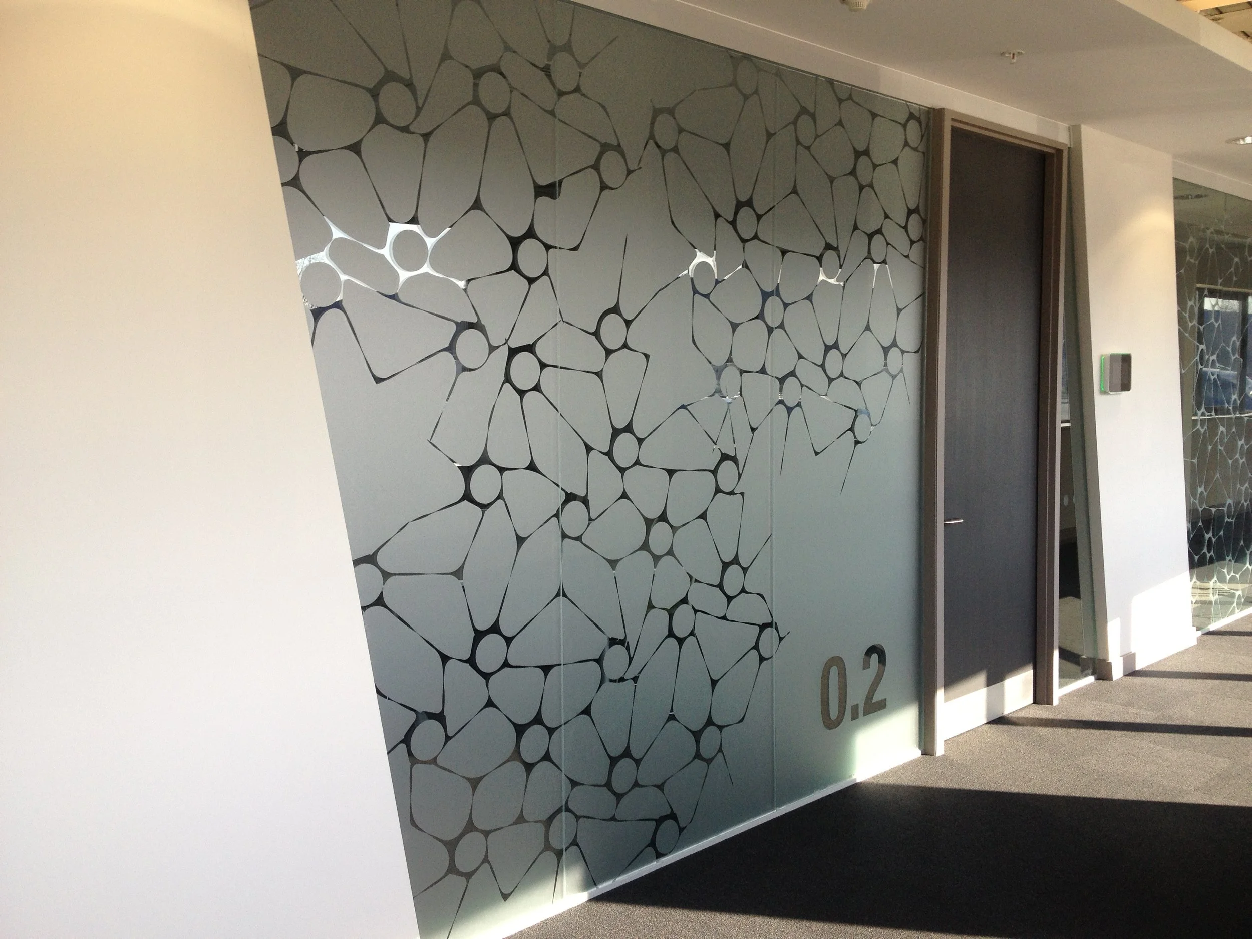 Modern interior with a wall featuring abstract black and white geometric patterns, a closed black door, and a small display or control panel on the wall. Sunlight casts shadows on the floor.