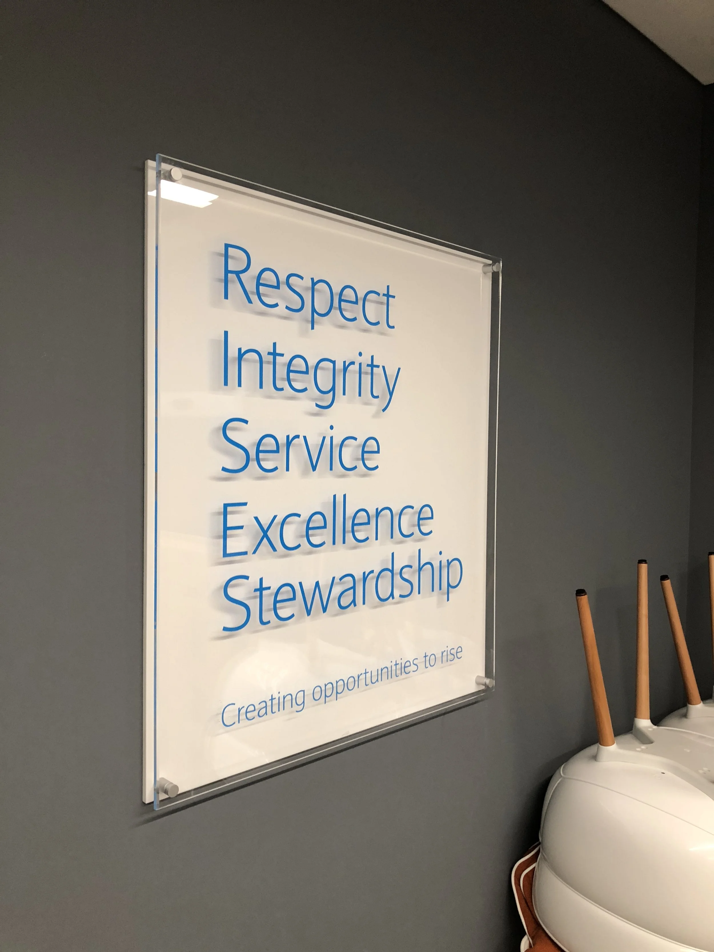 A white sign with blue text on a gray wall displaying core values: Respect, Integrity, Service, Excellence, Stewardship, with a subtitle 'Creating opportunities to rise'. To the right, parts of a white furniture piece with wooden legs are visible.