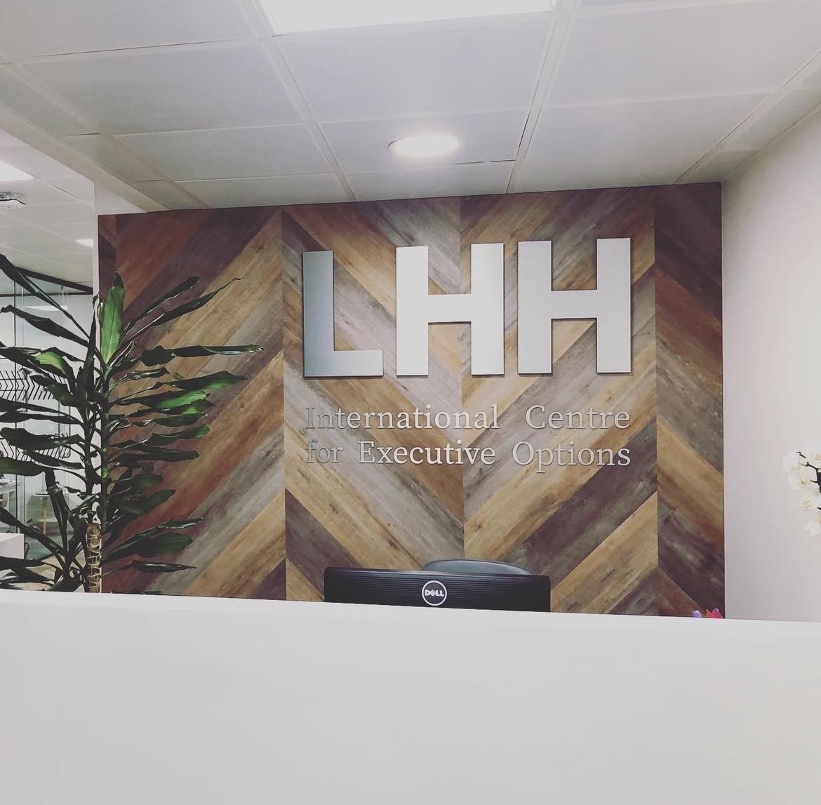 Reception area with a wall sign reading "LHH International Centre for Executive Options" with decorative wood paneling, a potted plant on the left, and a computer monitor on the reception desk.