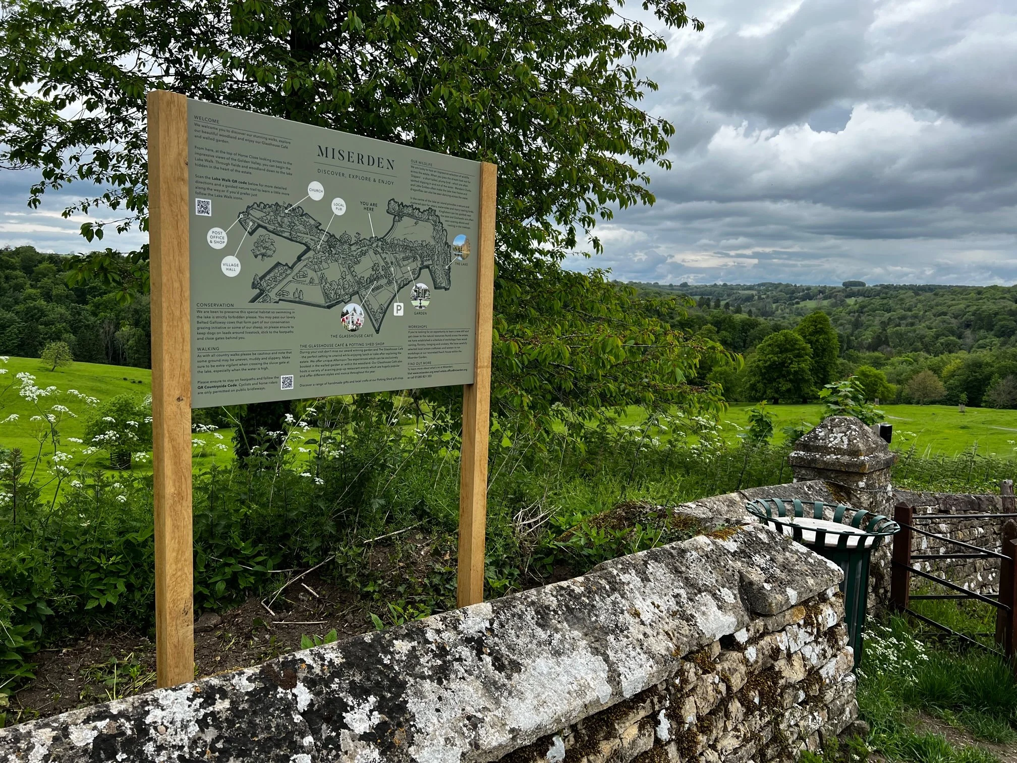 A landscape view of green fields with trees and a cloudy sky, featuring an informational signboard about Miserden. There is a stone wall, a trash bin, and a metal railing in the foreground.