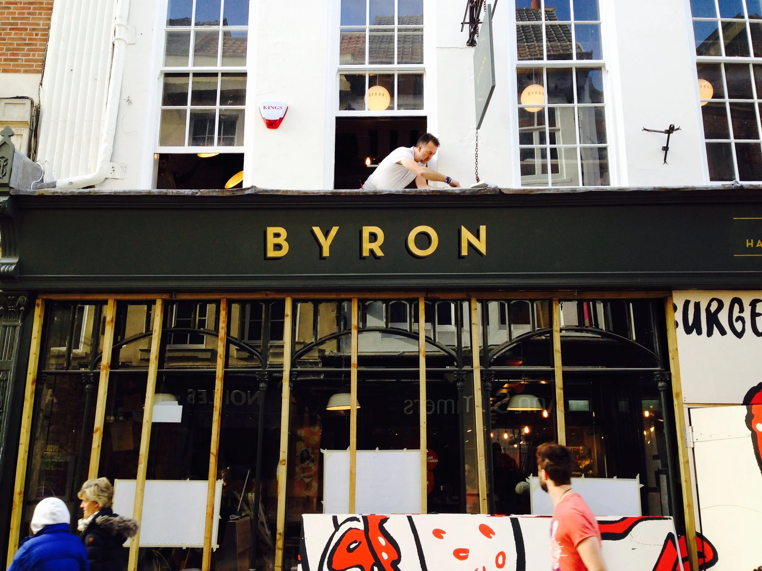 Exterior of Byron restaurant with a man working on the upper floor. Some people walking by in front with traditionally sign written gilded logo