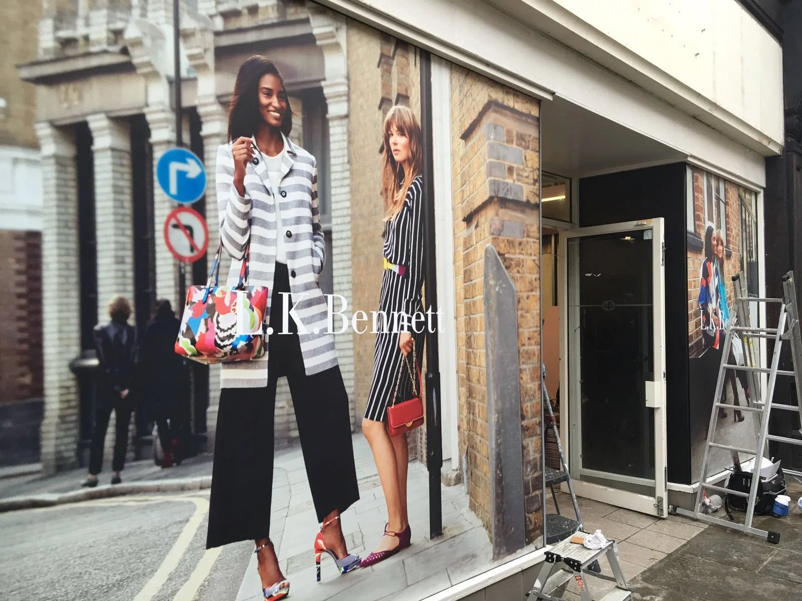 Store window display featuring large advertisements for fashion, showing women in stylish clothing and high heels on a city street.