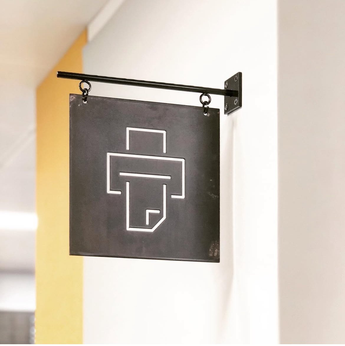 A black sign hanging from a metal rack on a white wall, featuring a minimalist outline of a first aid kit.