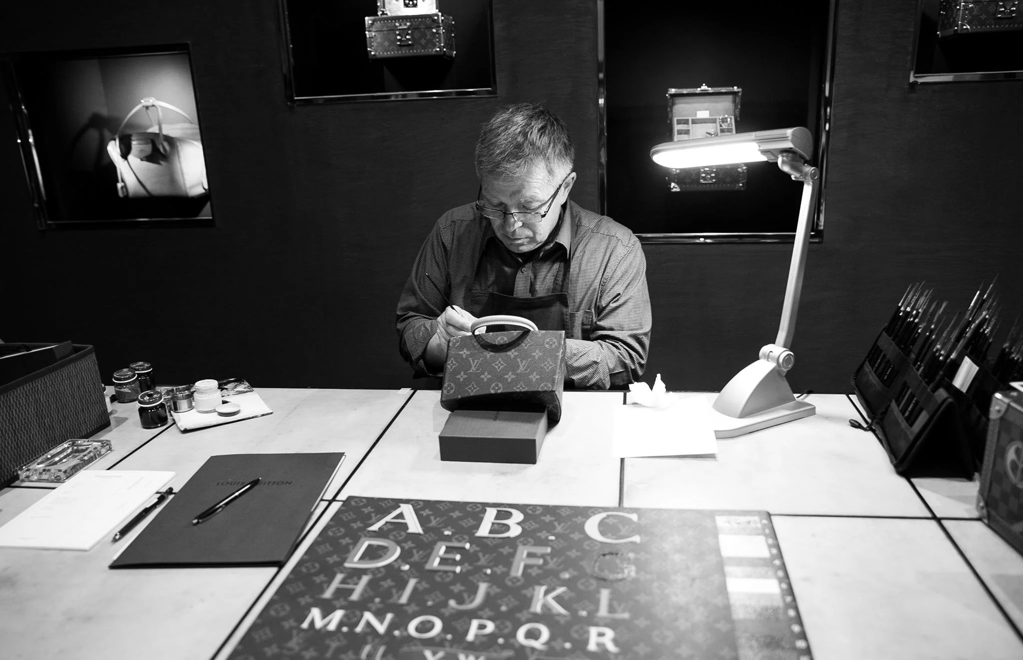 A man wearing glasses and an apron sits at a table, looking at a handbag under a dedicated lamp. The table has various items including small bottles, a Louis Vuitton folder, and a notebook. The background features dark walls with framed pictures and display cases.