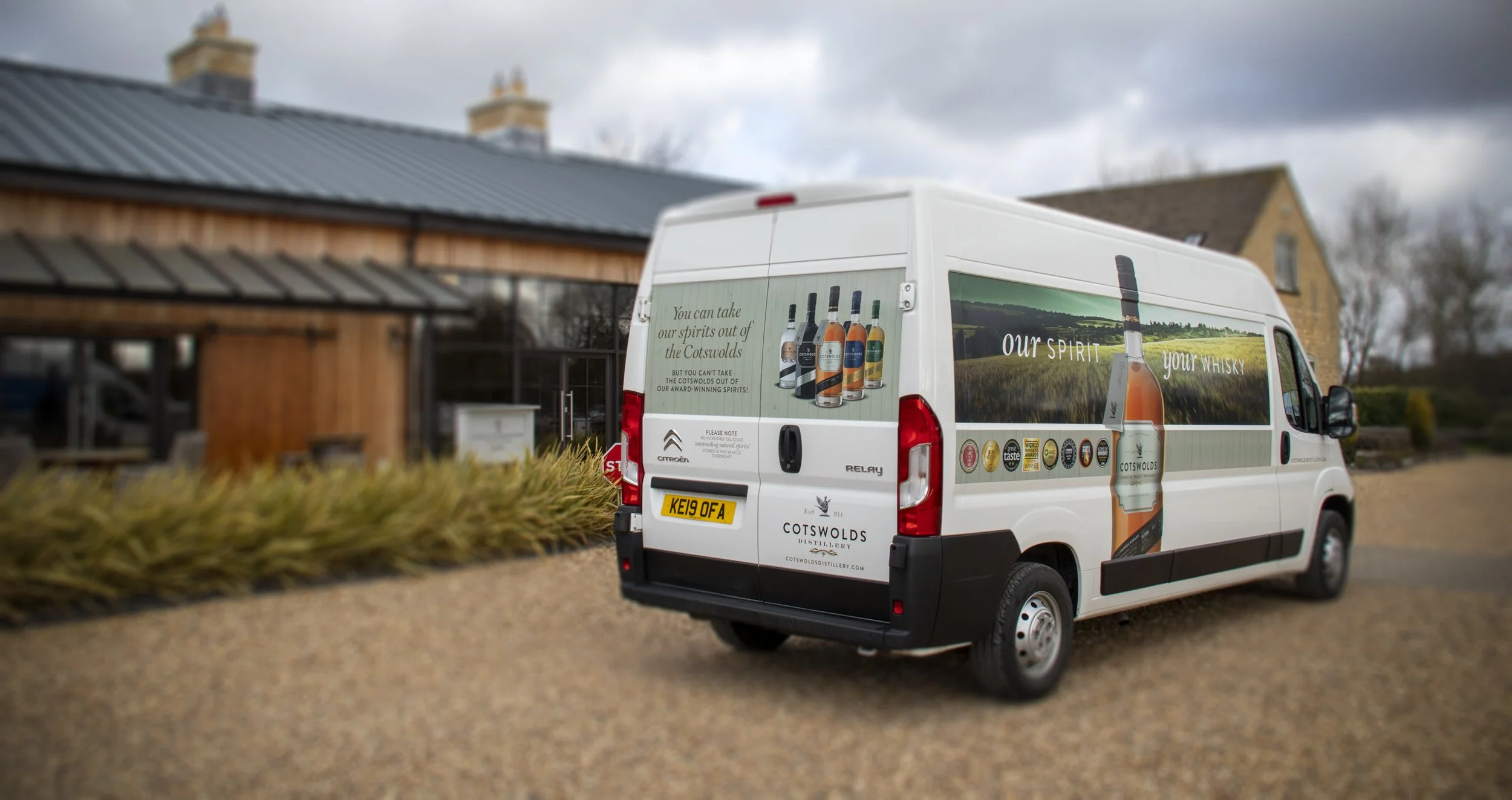 White delivery van with advertising for Cotswolds Distillery, featuring images of whisky bottles and slogans, parked on a gravel driveway near a wooden building with a metal roof and a brick house in the background.