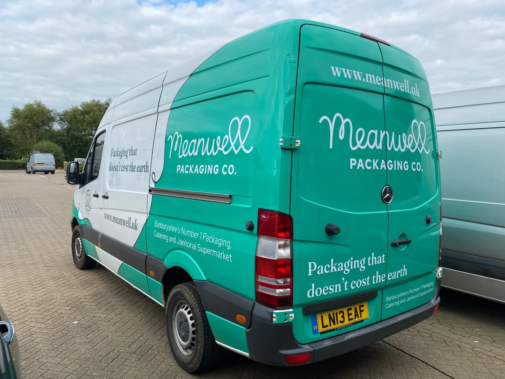 Green delivery van with the text "Meanwell Packaging Co." and slogan "Packaging that doesn’t cost the earth." parked on a paved surface, with other vehicles in the background.