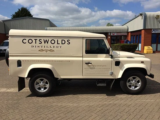White vehicle with Cotswolds Distillery branding parked in front of commercial buildings under cloudy sky.