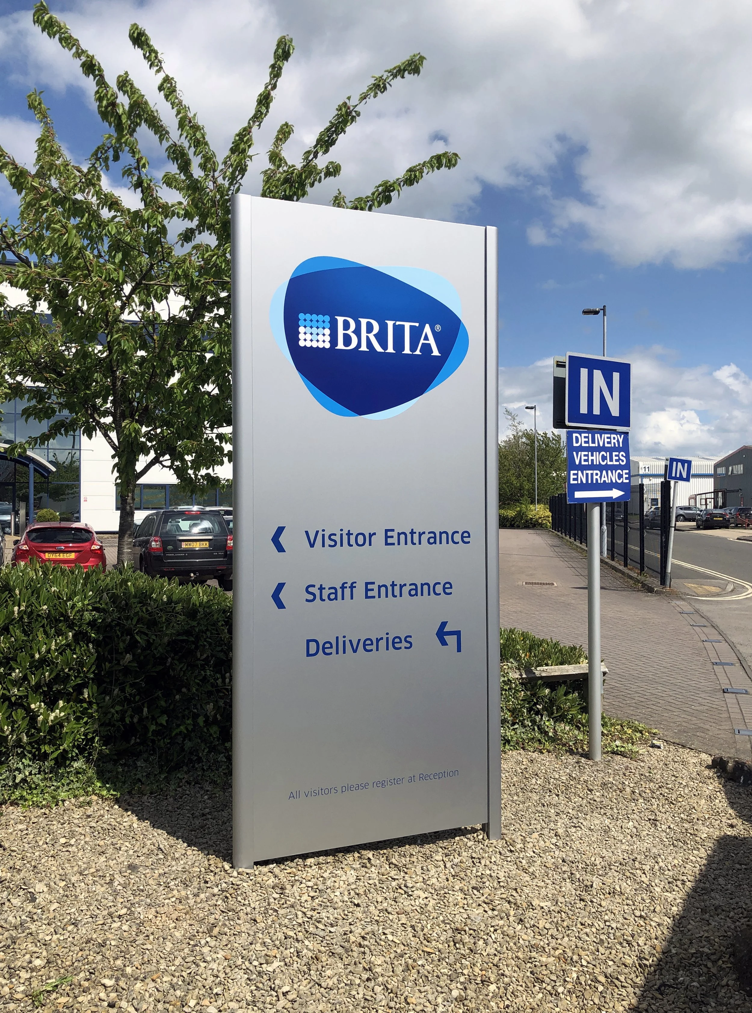 Sign outside a Brita facility indicating directions for visitor entrance, staff entrance, and deliveries, with an additional sign for delivery vehicle entrance. The sign is next to a sidewalk, parked cars, and a tree under a partly cloudy sky.