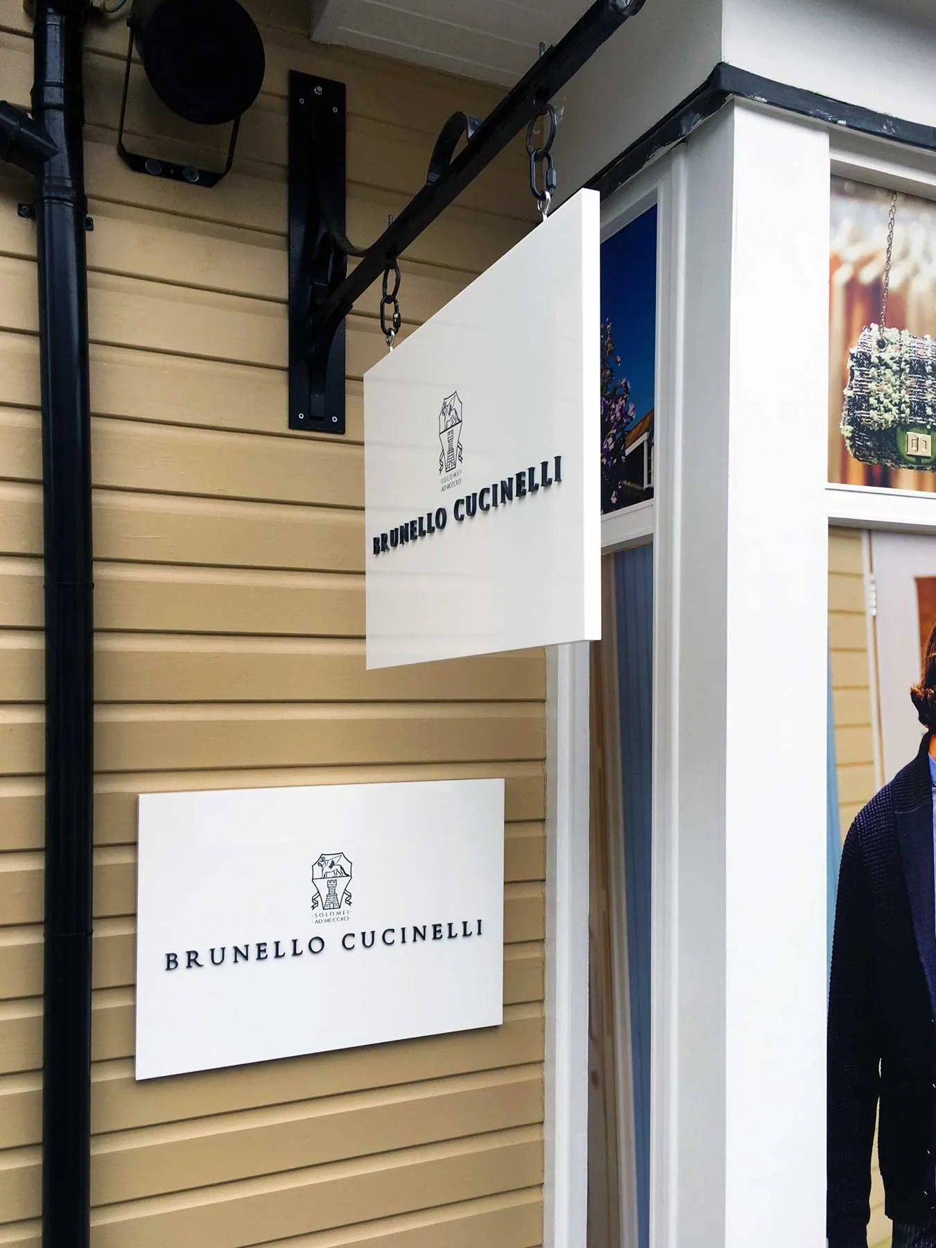 Storefront signage for Brunello Cucinelli, with the logo and name displayed on a hanging sign and a wall-mounted sign.