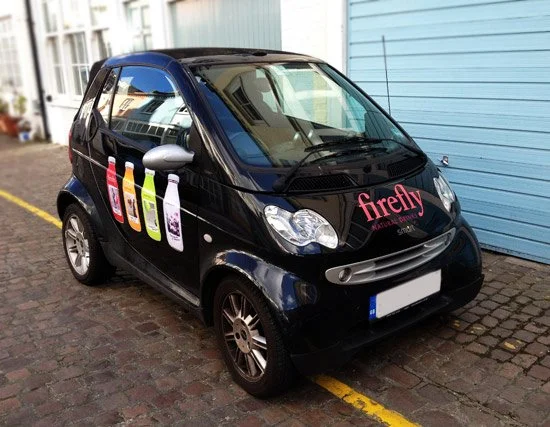 Black Smart car parked on a cobblestone street with a blue building in the background, featuring colorful Firefly cleaning product bottles and the Firefly logo on the hood.