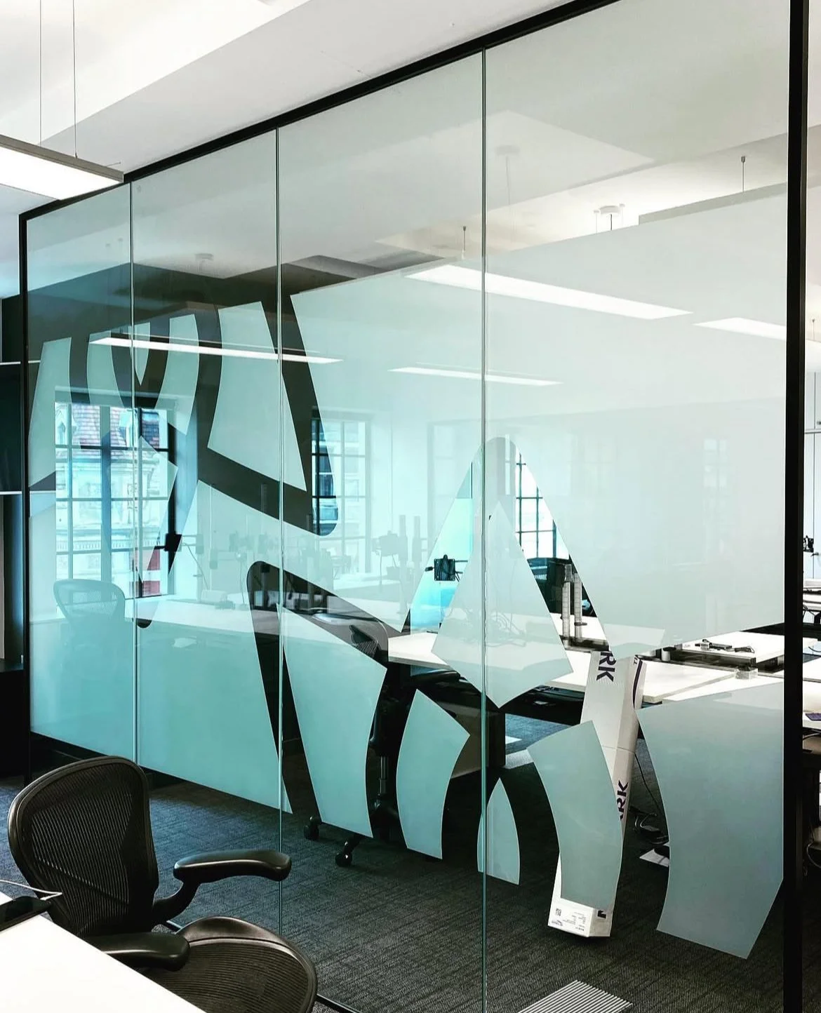 Modern office with glass wall featuring large black and white logo or design, desks, chairs, and office equipment visible through the glass.