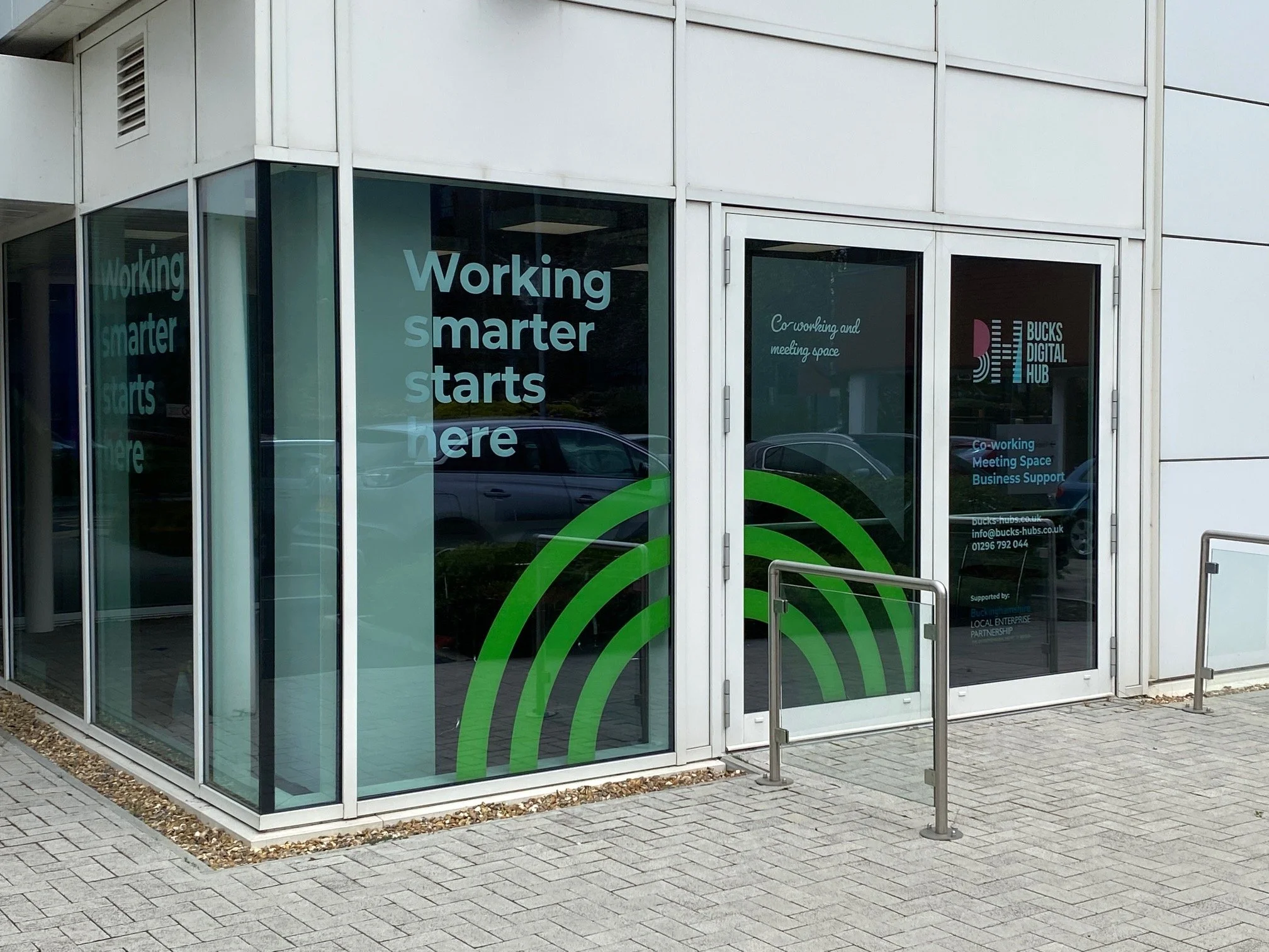 Exterior of a modern office building with glass walls and doors, featuring signs that say 'Working smarter starts here,' and a logo for Bucks Digital Hub, with parking and reflection of cars outside.