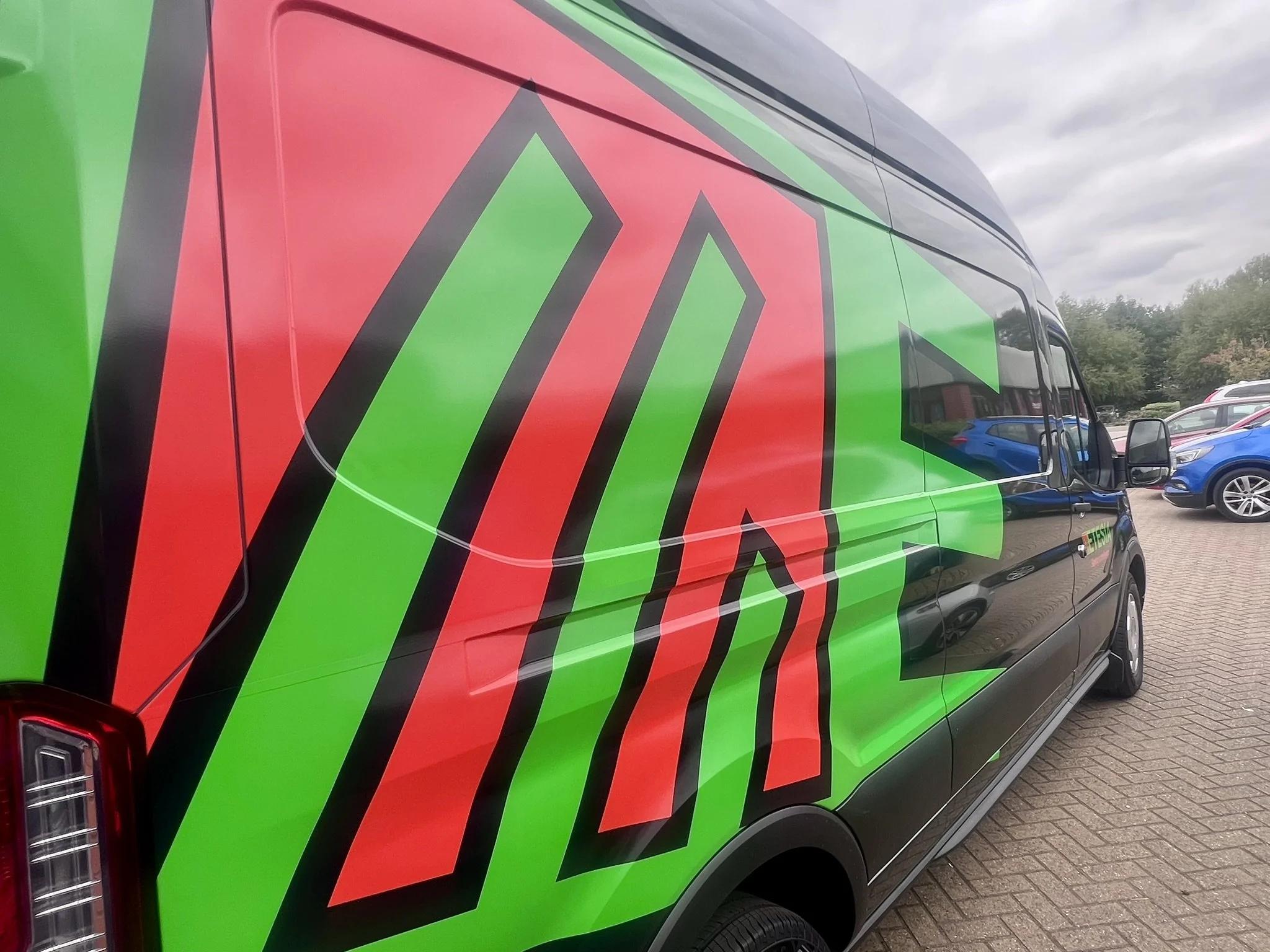 A black van with a large green and red design on its side, parked on a paved lot with other vehicles and trees in the background.
