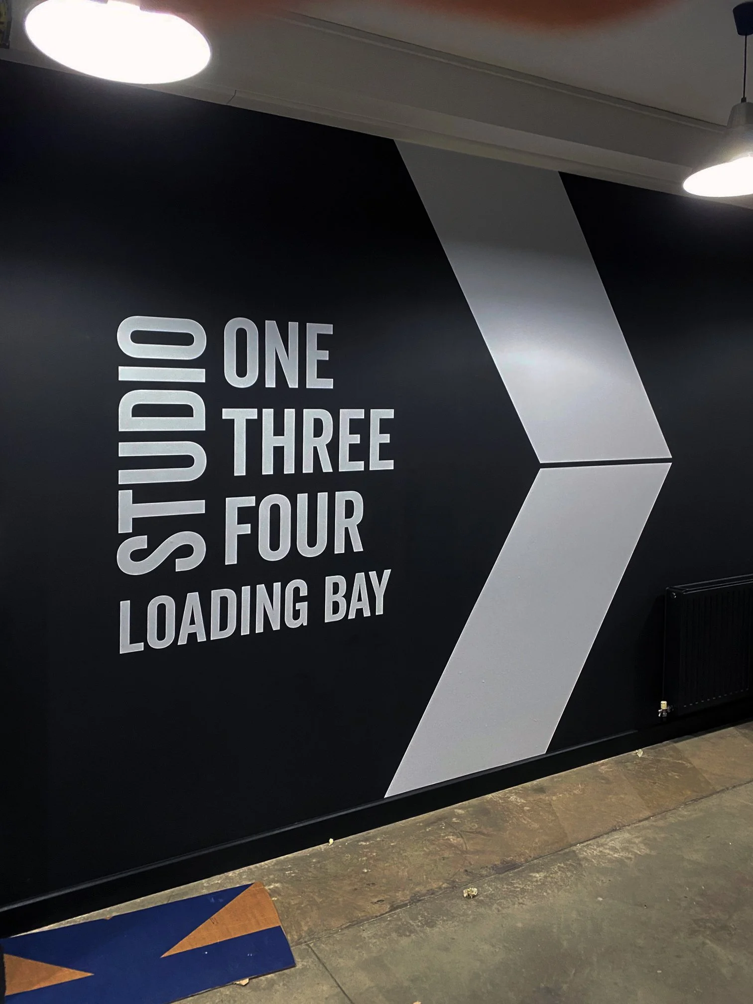 A wall with military-style instructions in white letters on a black background, showing steps for a process: "STUD 0, ONE, 1, THREE, 2, FOUR, 3, LOADING BAY." There are lights overhead and a section of the floor visible in the foreground.