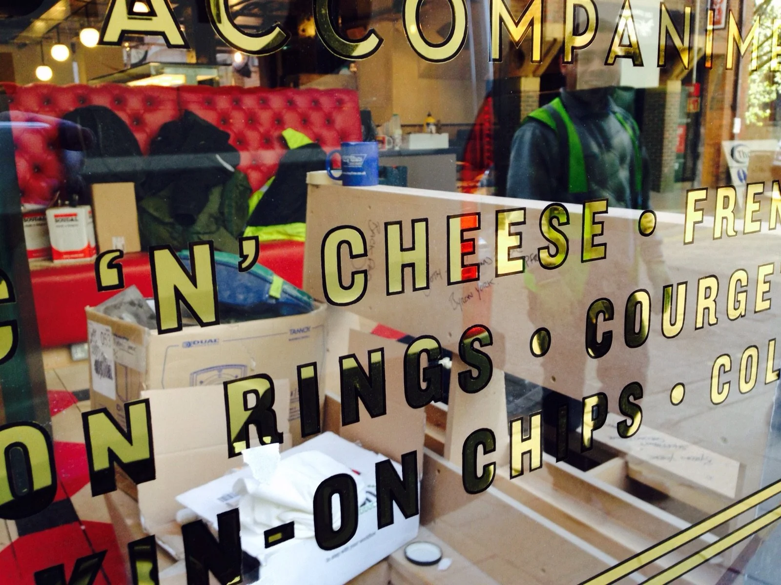 View through a glass window of a restaurant or cafe showing interior items such as a red upholstered booth, boxes, and clothing. The window has yellow text listing food items like cheese, fried, wings, chips, and crown.