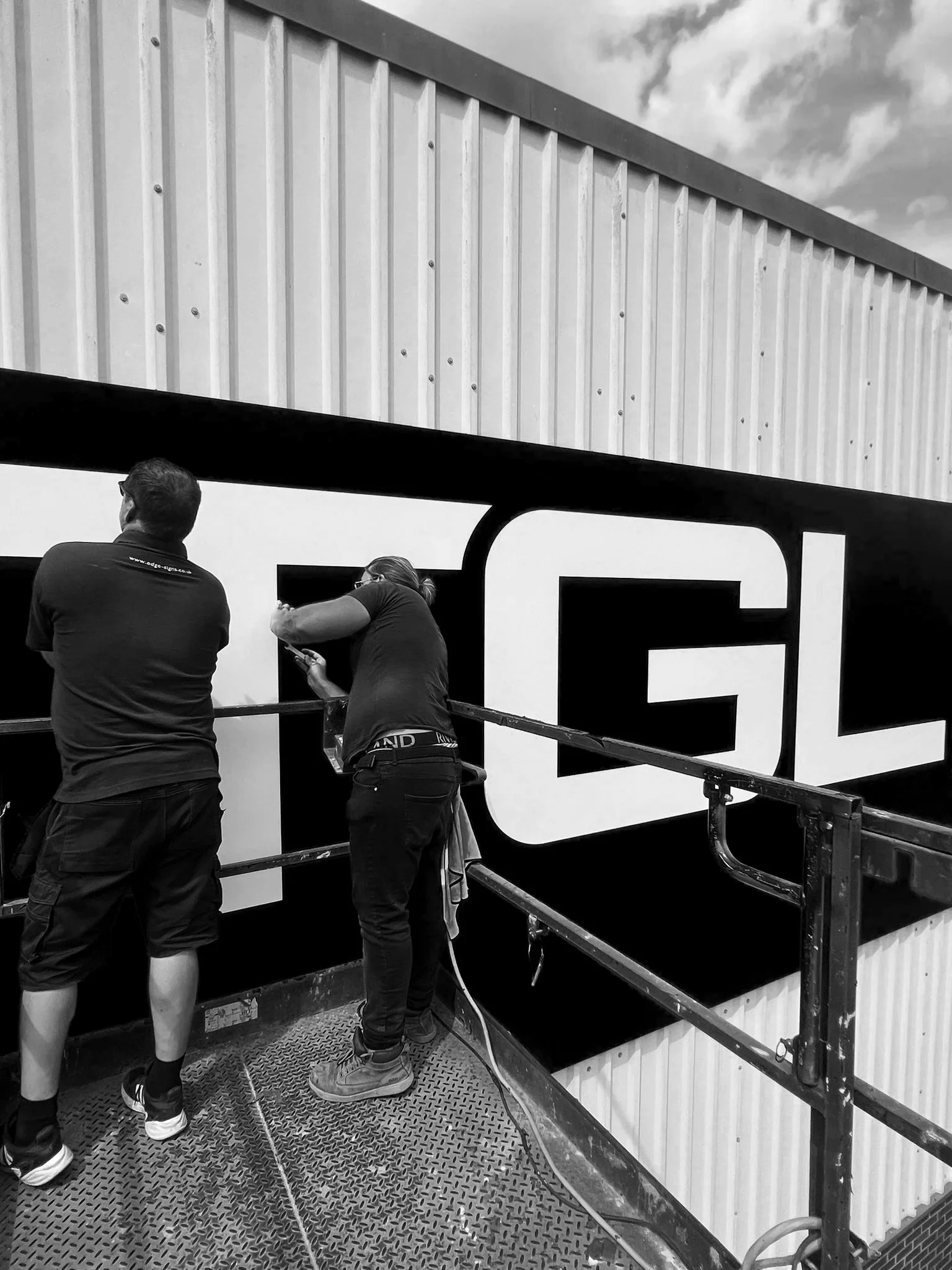 Two workers painting a large sign on the side of a building, with a metal railing platform and a cloud-filled sky in the background.